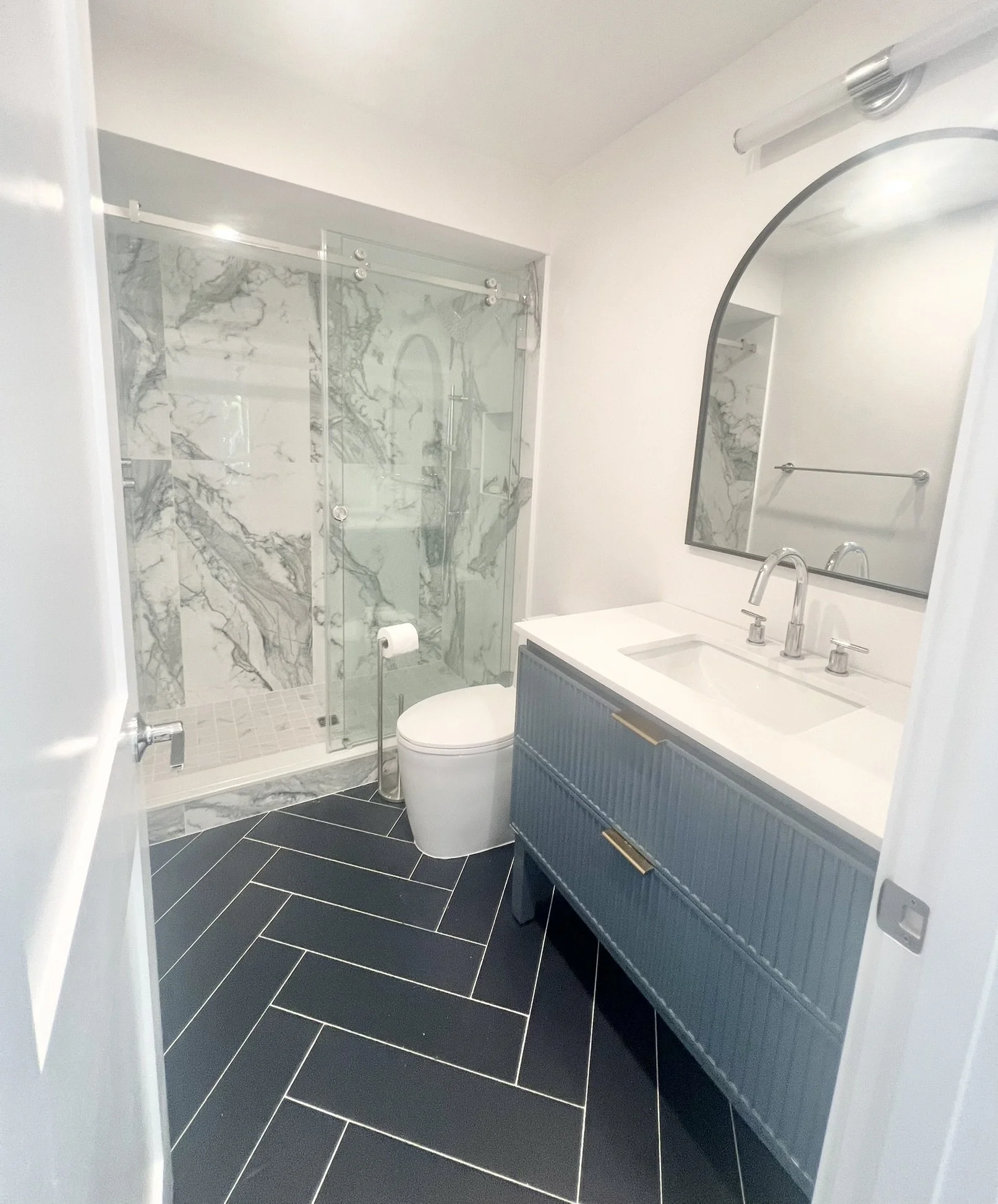 Modern bathroom with a glass shower stall, a white toilet, and a blue vanity with a white countertop and sink. A large mirror is mounted above the sink, and the floor has dark herringbone tiles.