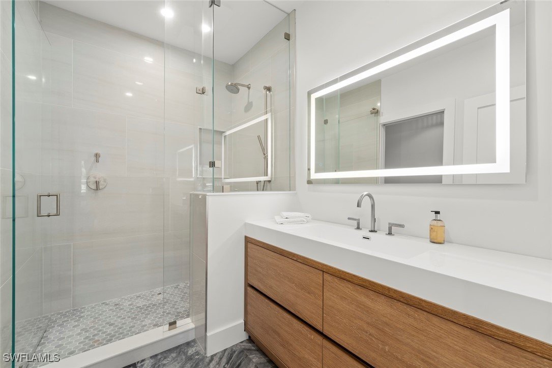 Modern bathroom with a glass-enclosed shower, a large backlit rectangular mirror, wooden vanity, and white walls.