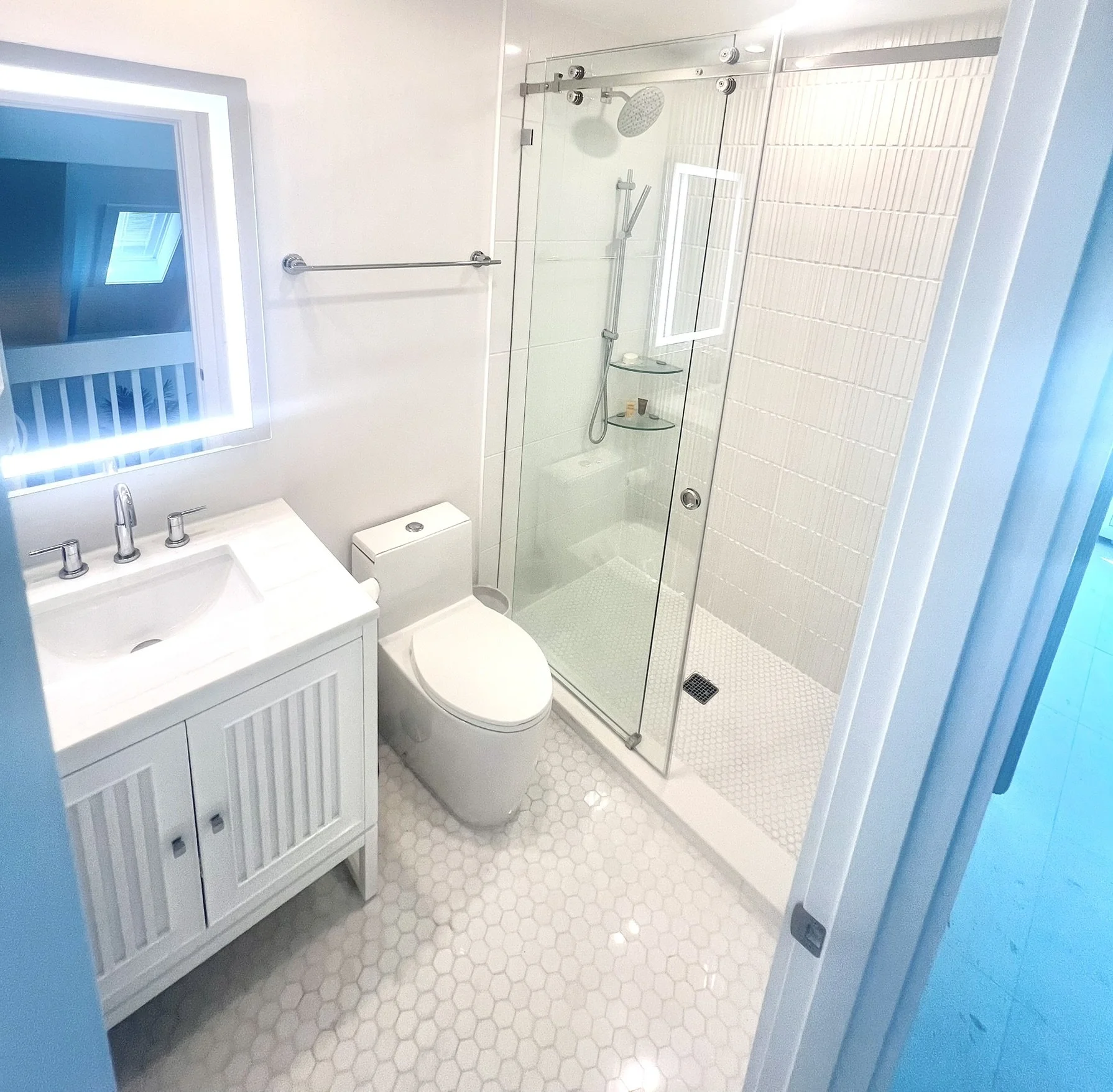Small modern bathroom with a white vanity and sink, toilet, and a glass-enclosed shower with a rain showerhead. White hexagonal floor tiles, a mirror, and natural light from a window.