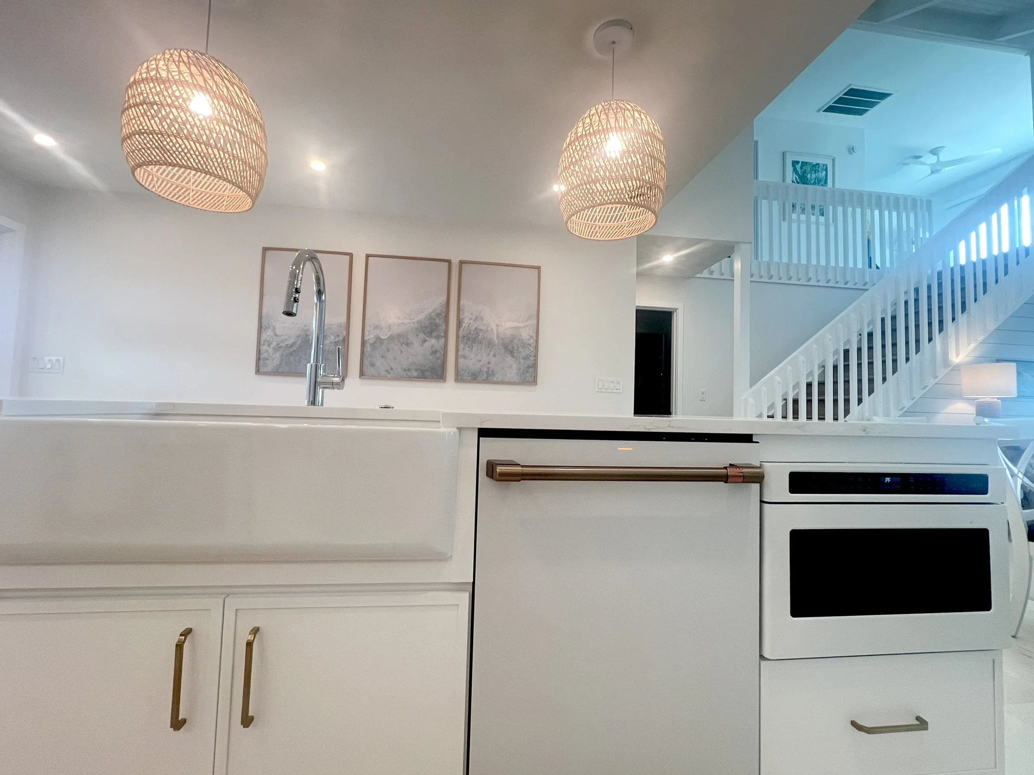 Modern kitchen with white cabinets, a large sink, and a brass handle on the dishwasher. Wooden pendant lights hang from the ceiling. In the background, a staircase with white railings and a hallway with artwork and a ceiling fan.