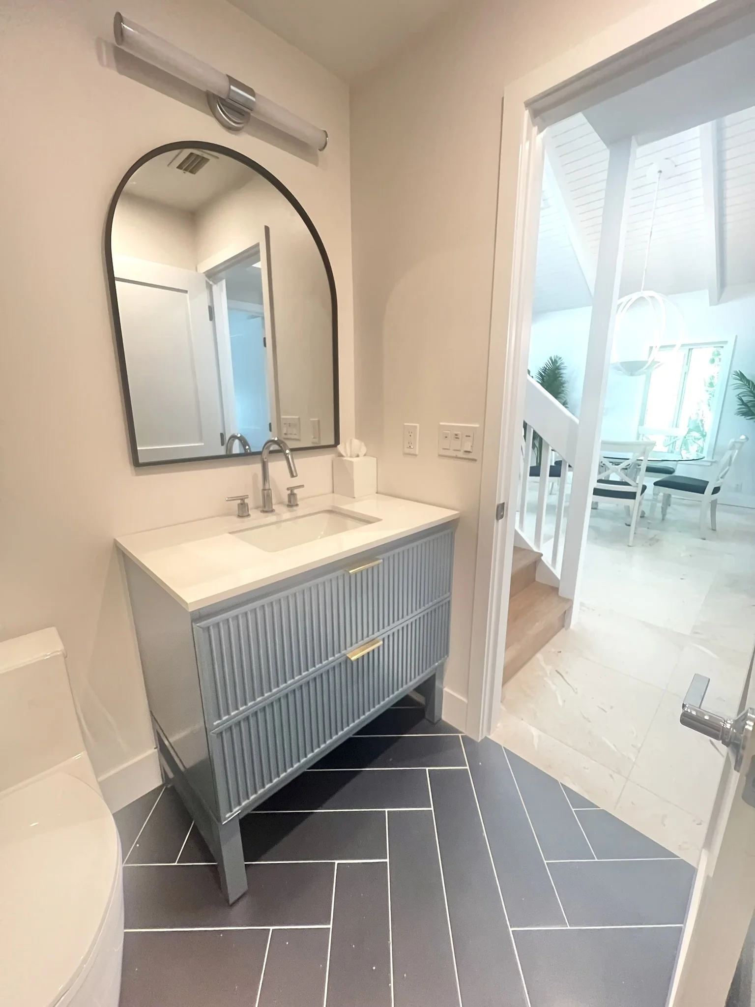 Modern bathroom with black tile flooring, a gray vanity with ribbed detailing, a mirror, and a white countertop with a built-in sink. There is a tissue box on the vanity, a white toilet to the left, and a doorway leading to a bright room with white f