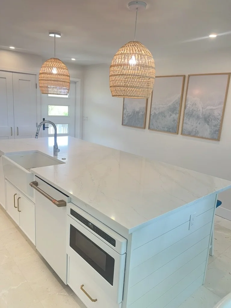 Modern kitchen with white island, marble countertop, built-in oven, and wicker pendant lights hanging from ceiling. Framed mountain artwork on the wall.