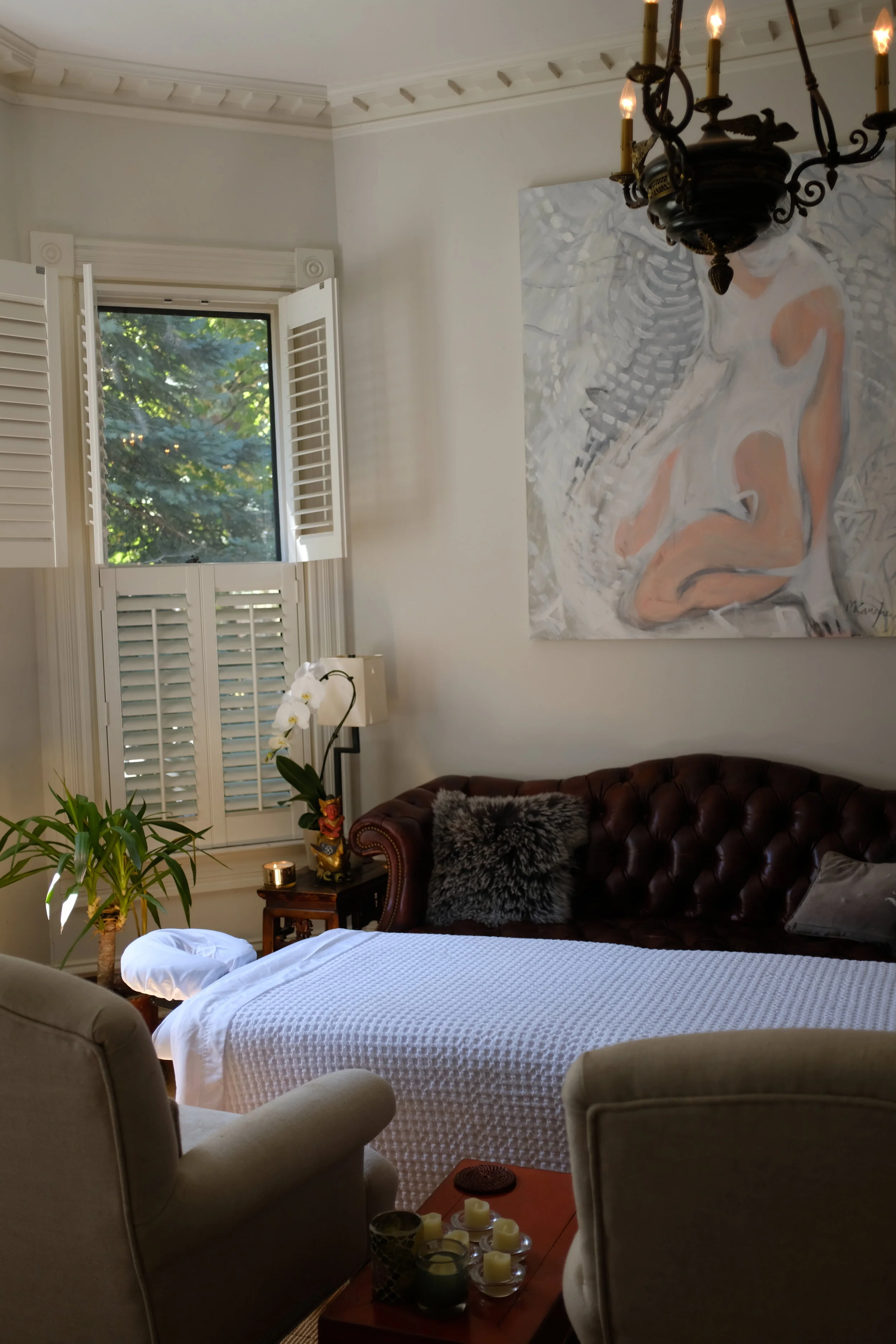 Living room with a window open to green trees, a large abstract nude painting on the wall, a vintage chandelier hanging from the ceiling, a brown leather sofa with a textured blanket and a furry pillow, a small wooden side table with candles, and some houseplants near the window.