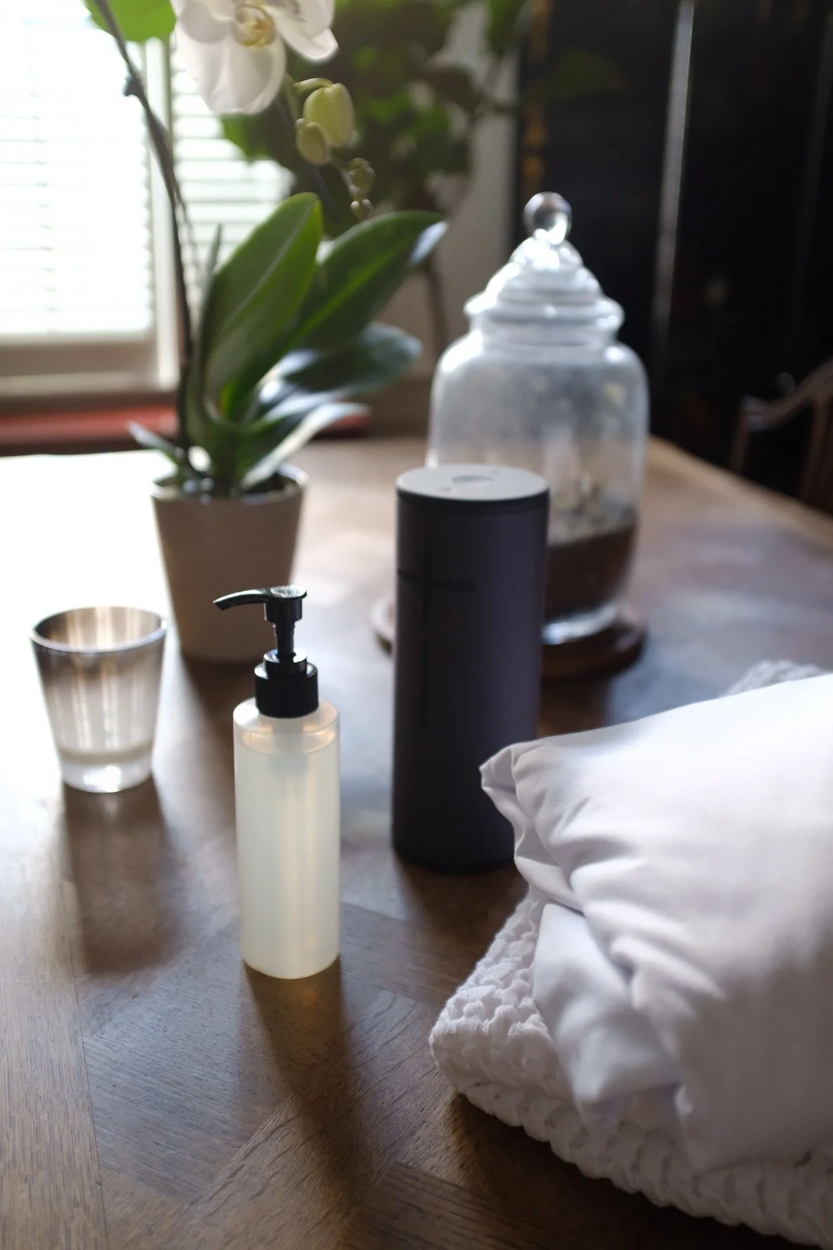 A table with a potted orchid plant, a small clear cup, a white pump soap dispenser, a tall black container, a glass jar with a lid, a white cloth, and a pillow, with a window and blinds in the background.