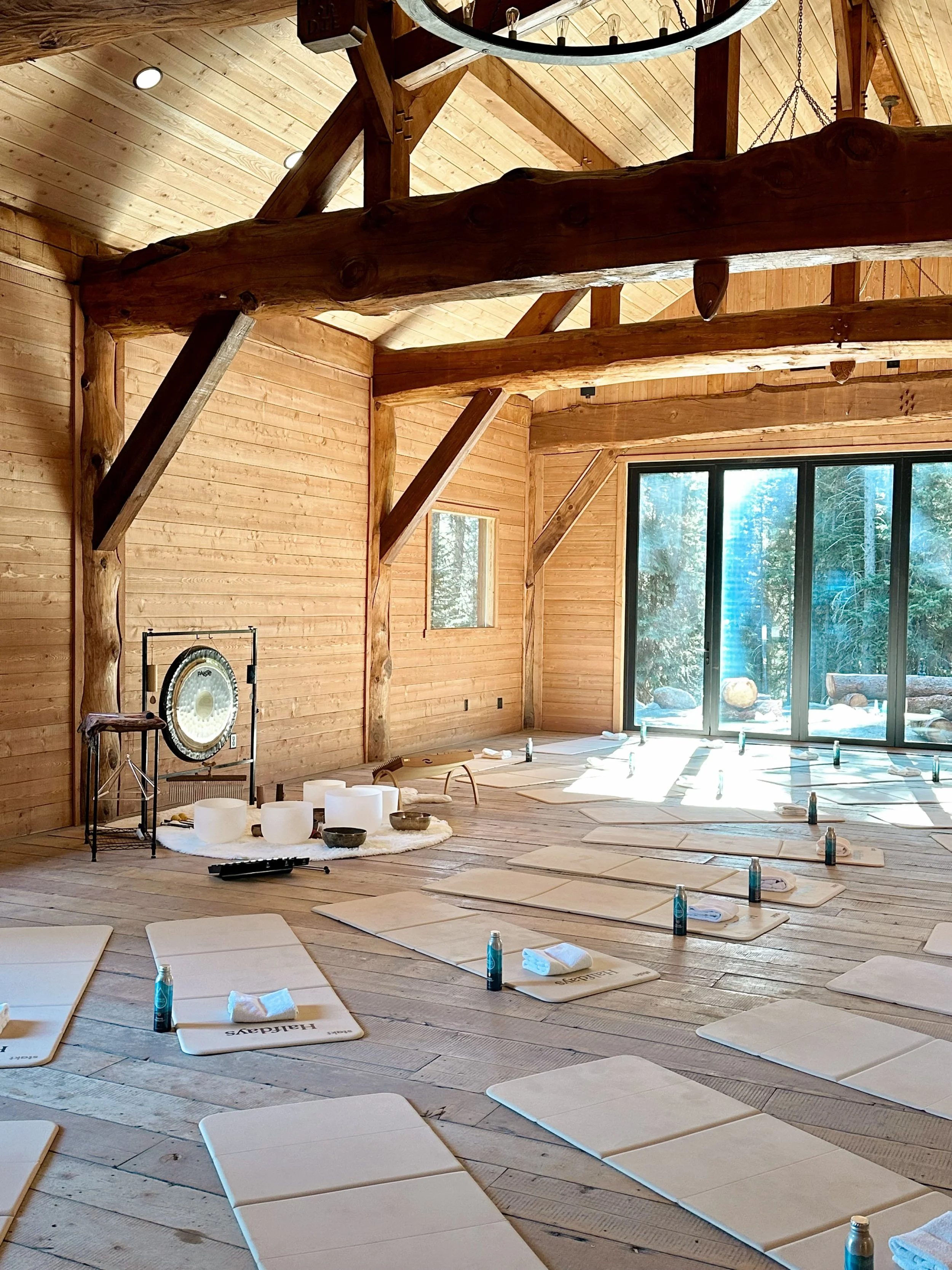 A spacious wooden room with large sliding glass doors, a high vaulted ceiling with wooden beams, and a window showing a forest. The floor is set with meditation mats, water bottles, towels, and small bowls. There are crystal singing bowls and a gong in the corner, suggesting a meditation or yoga class setup.