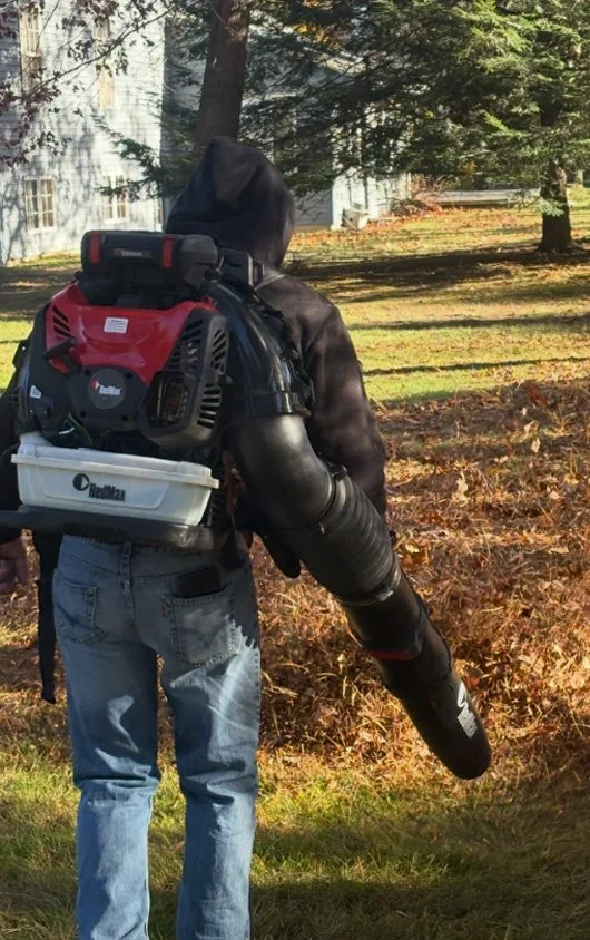Person in a black hoodie and blue jeans carrying leaf blower outdoors on a sunny day, with trees and a house in the background.