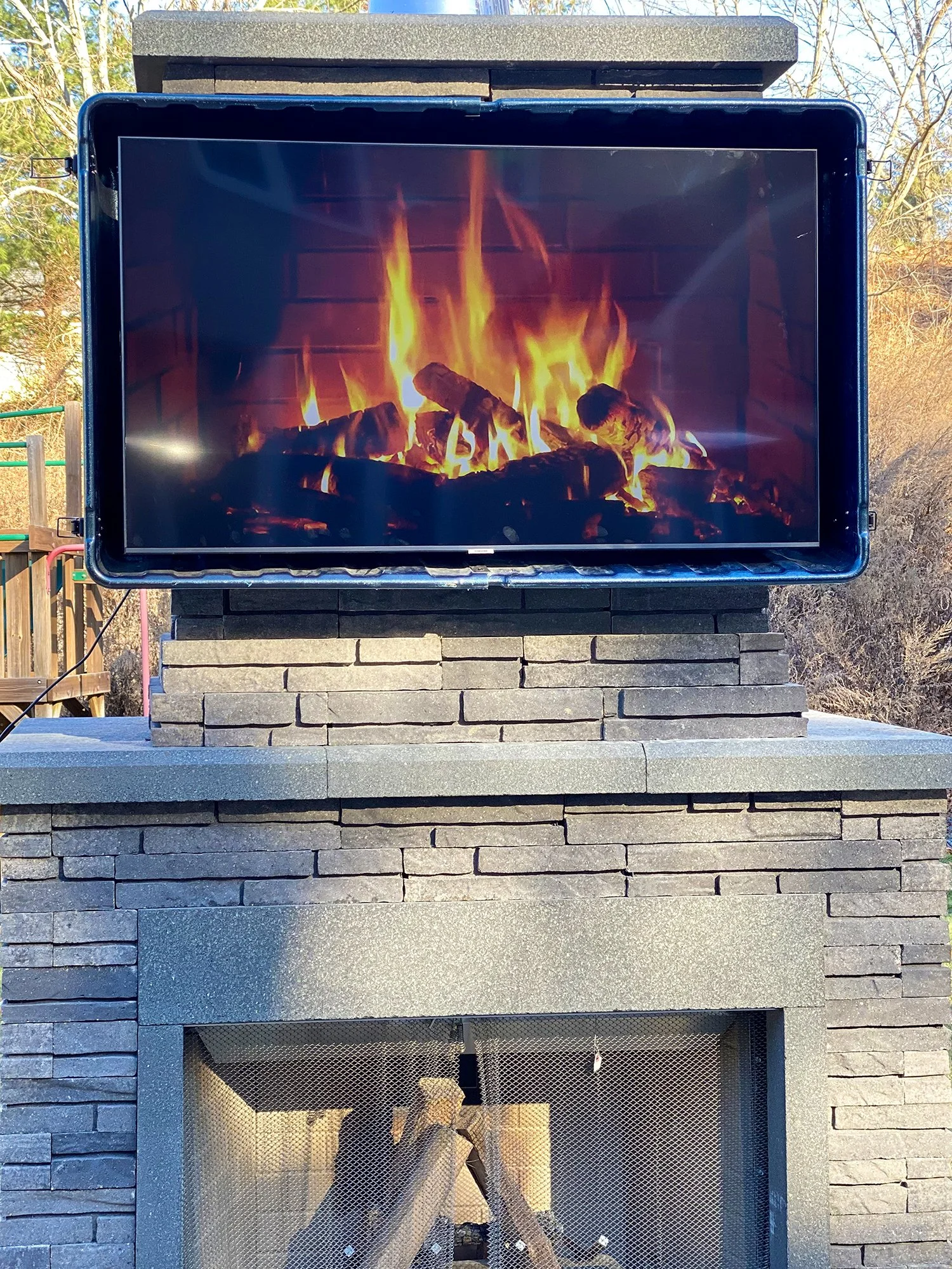 A screen displaying an image of a burning fireplace with logs, mounted above an outdoor fireplace or firepit made of dark brick stone.