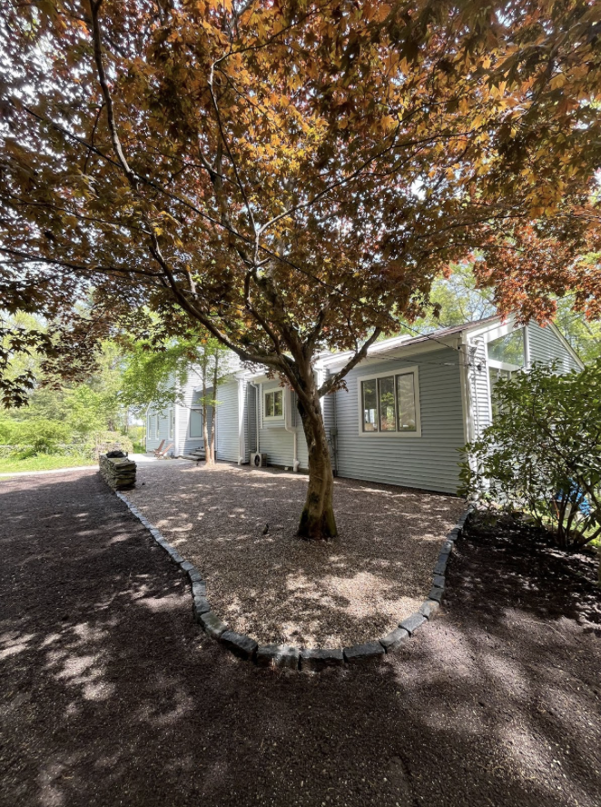 A house with gray siding surrounded by a landscaped yard with a large tree, a gravel pathway, and a stone border.