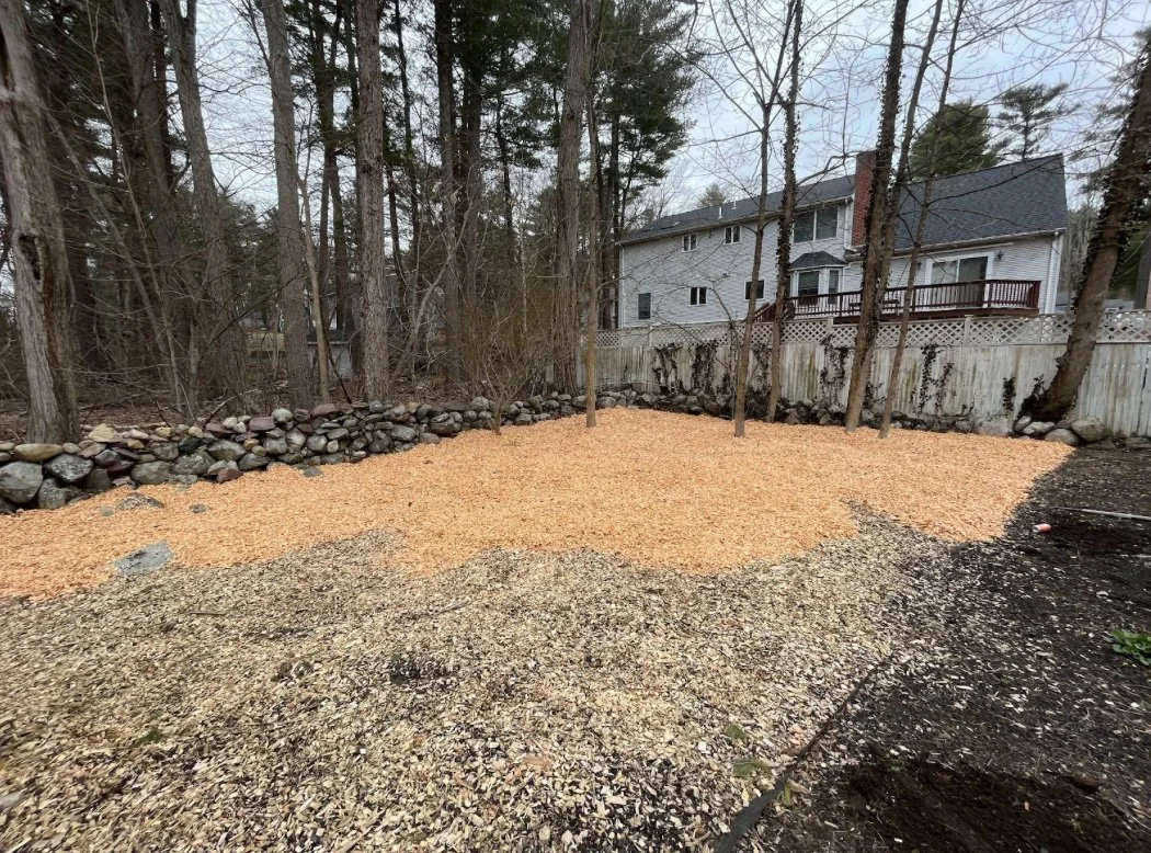 Backyard with a gravel patch, a retaining wall made of rocks, and a wooden fence with trees in the background.