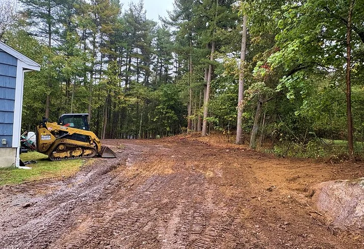 A dirt driveway or path in a wooded area with a small yellow excavator parked on the left near a blue building.