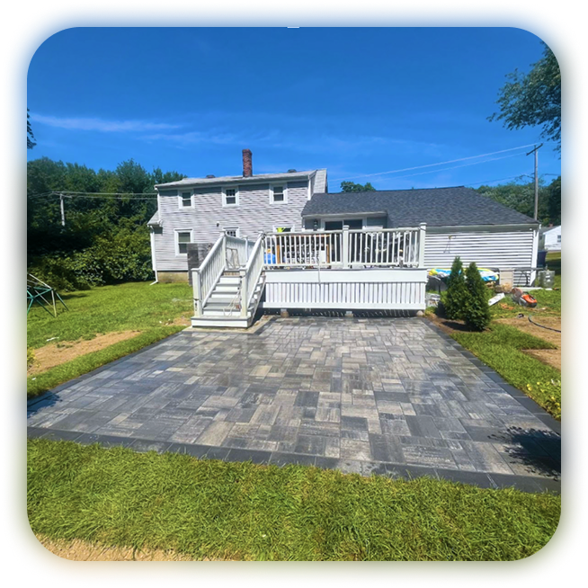 Backyard with a newly installed paved patio, white deck with stairs, grassy lawn, and a house with a chimney under a clear blue sky.