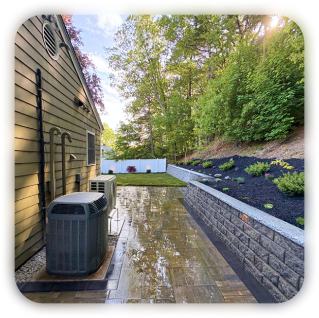 Backyard with a wet stamped concrete patio, green grass, a white fence, and a raised garden bed with plants, bordered by trees.