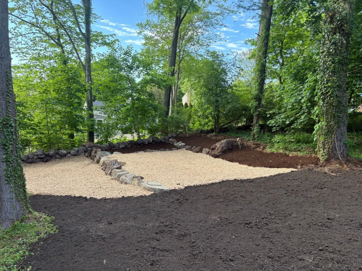 A backyard scene under a cloudy sky, with a newly landscaped area featuring gravel surrounded by rocks, and dark soil around it, with tall trees and greenery in the background.