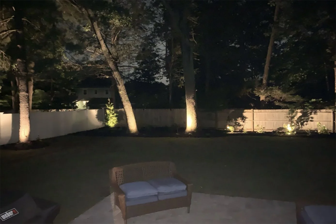 Nighttime backyard scene with trees, a wooden fence, and outdoor lighting illuminating the trees and plants. A patio area with a wicker bench and outdoor furniture.