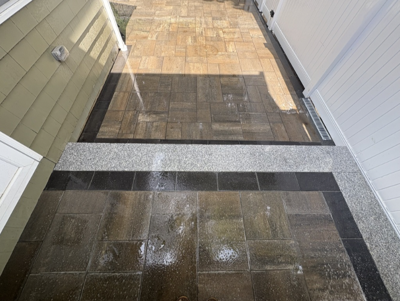 Wet outdoor patio area with brown and black tiles and a gray granite strip, located next to a green house wall and a white fencing.