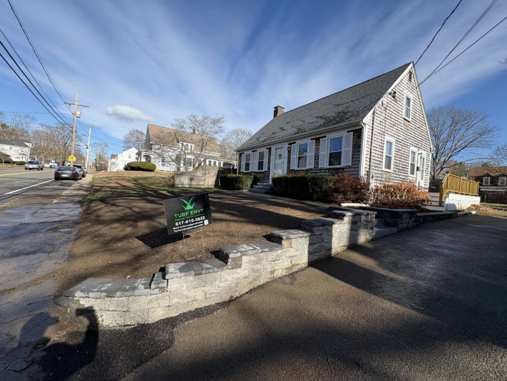 Residential house with a sign in the yard reading 'Turf Envy' and a phone number, with a newly built stone retaining wall in the front yard and a clear blue sky above.
