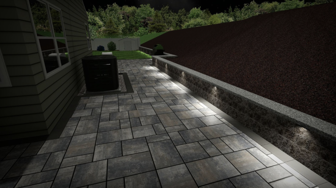 A backyard patio with stone pavers, outdoor lighting, and a low wall. There is grass, trees, and a shed in the background.