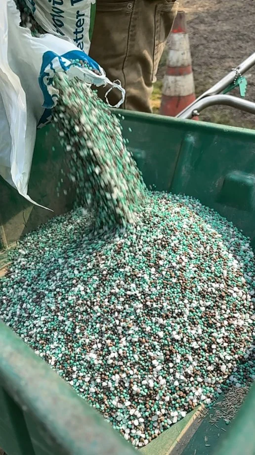 People pouring a mix of small, colorful pellets into a large green container outdoors, with construction cones and a shovel nearby.