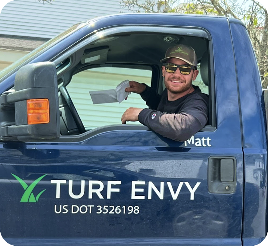 A man sitting in the driver's seat of a blue Turf Envy truck, smiling and holding a napkin. He is wearing sunglasses, a cap, and a long sleeve shirt. The truck has the name Matt on the door and a logo with green grass blades.