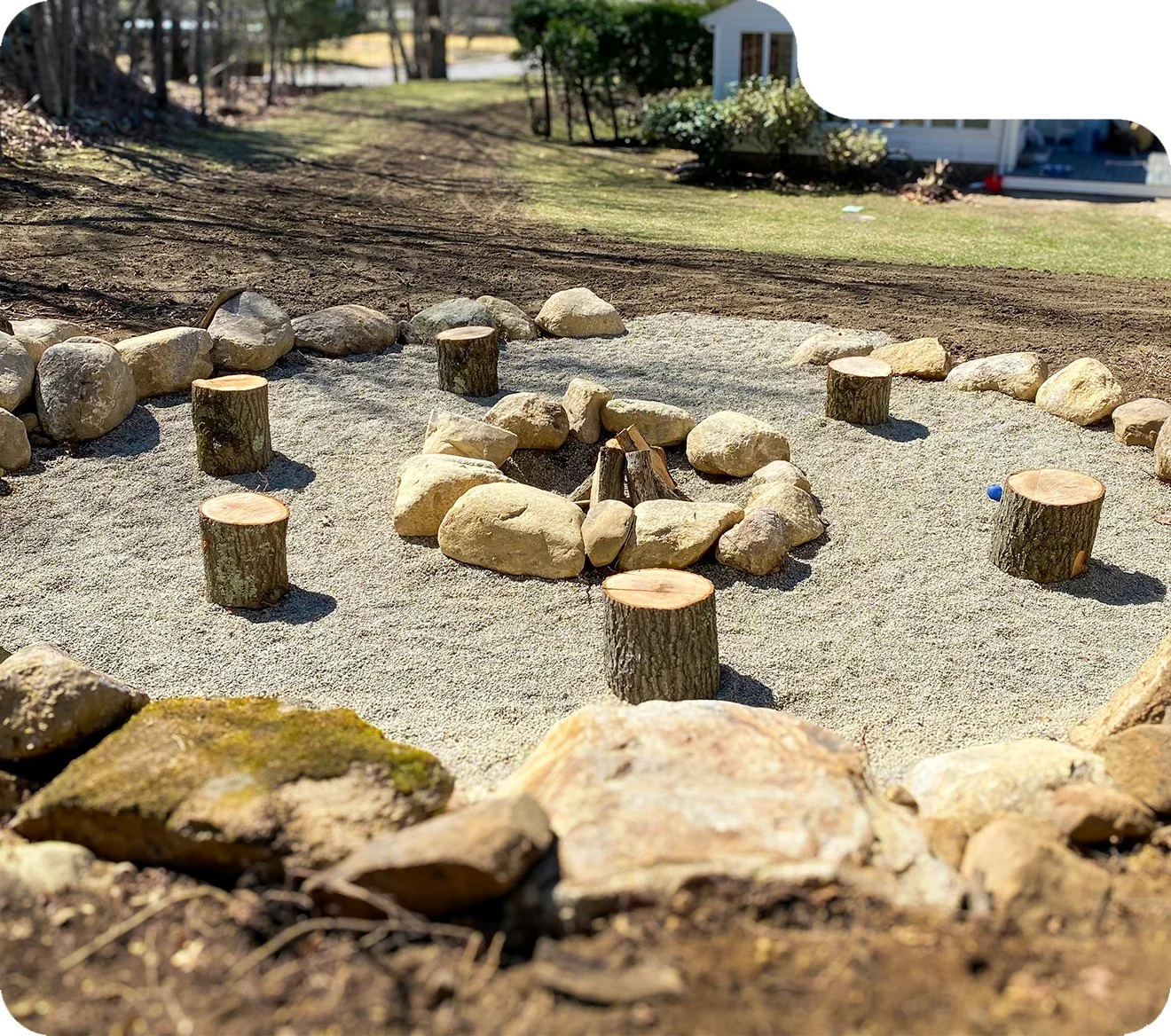 A circular fire pit area made of rocks and small tree stumps on sand, outdoors in a backyard with grass, trees, and a white house in the background.