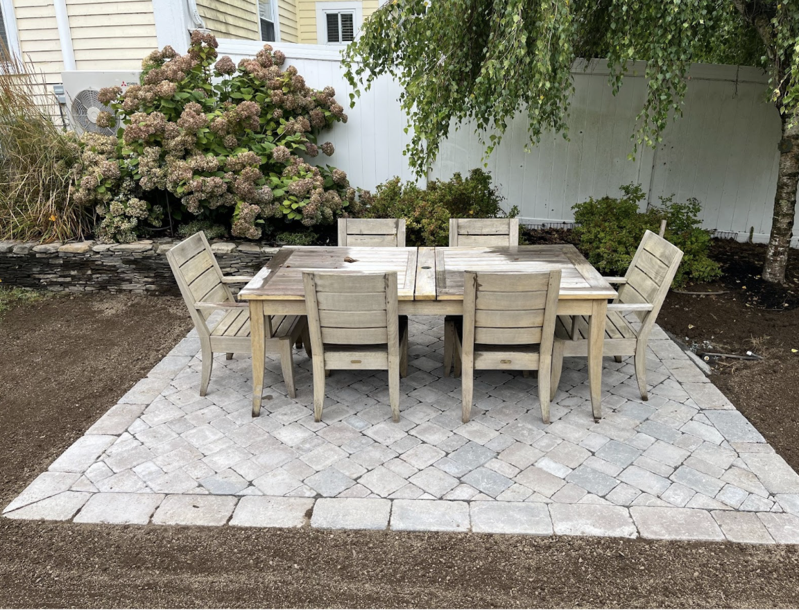 Outdoor patio with a wooden table and six matching chairs on a stone-paved surface, surrounded by flowering bushes and trees with a white fence in the background.