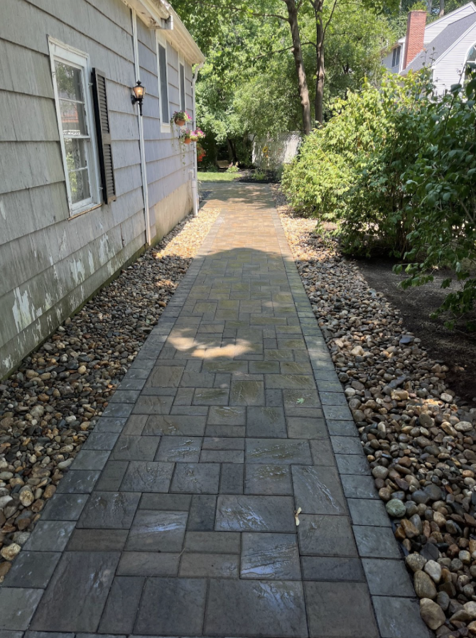 A paved walkway with interlocking stone bricks runs alongside a house with exterior siding, black shuttered windows, and decorative hanging flower baskets. The walkway is bordered by small rocks and green bushes, leading to a shaded yard with trees in the background.