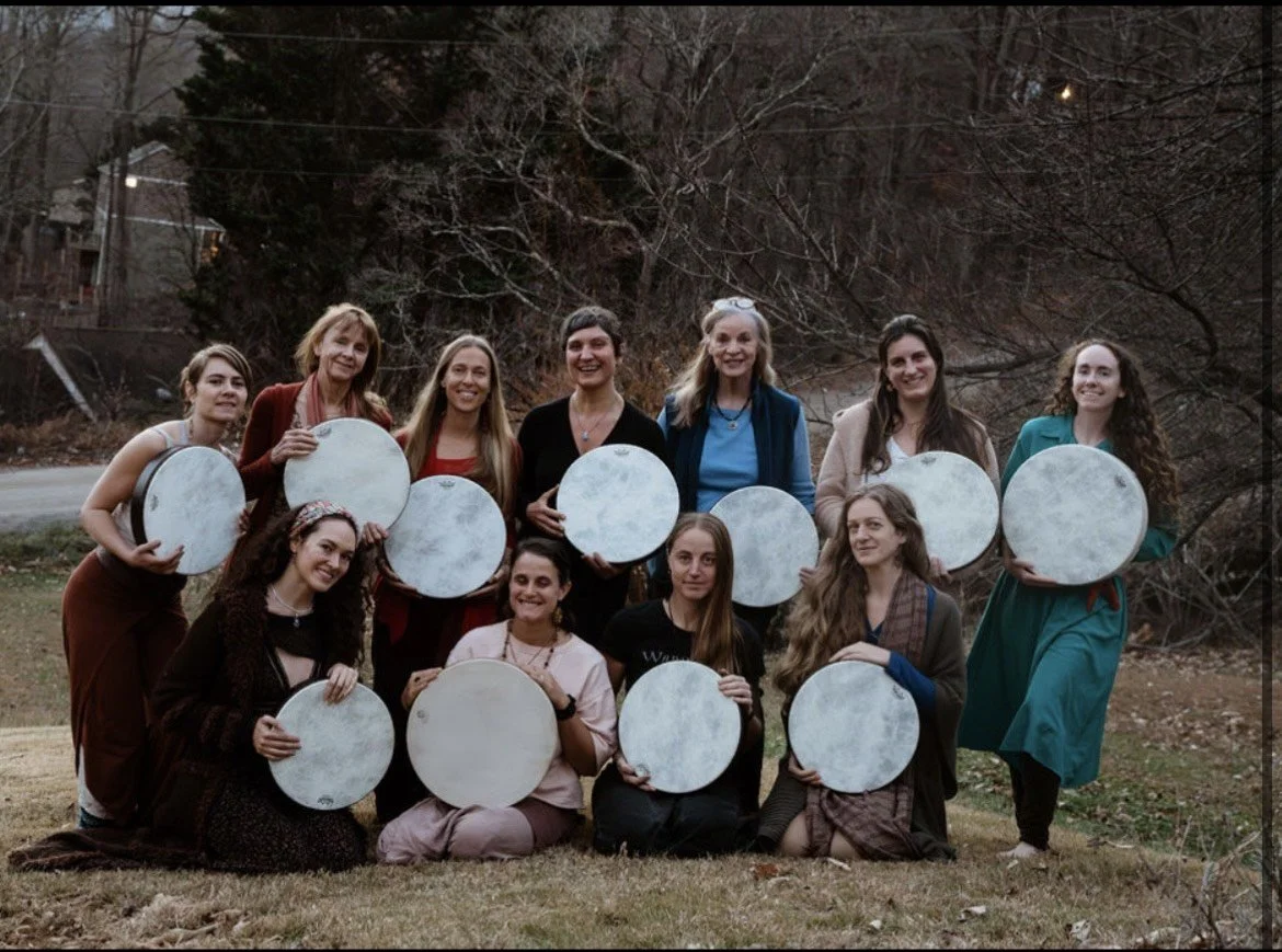 Group of women and girls outdoors holding tambourines during daytime, autumn trees in background.