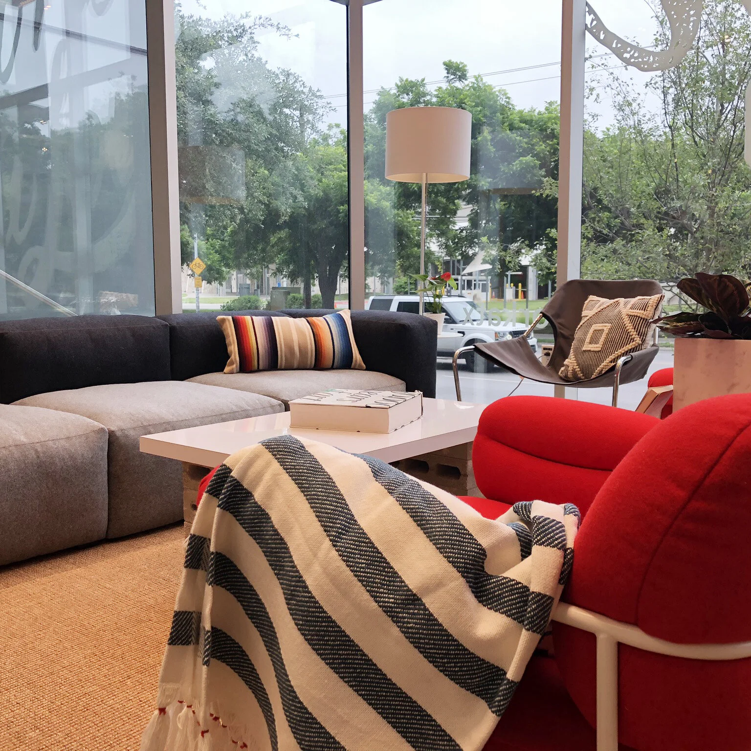 A cozy waiting area with a gray sofa decorated with multi-colored striped pillows, a red armchair with a striped throw blanket, a white coffee table with a box on it, a tall white floor lamp, a black chair with a cushion, and large glass windows showing trees and parked cars outside.
