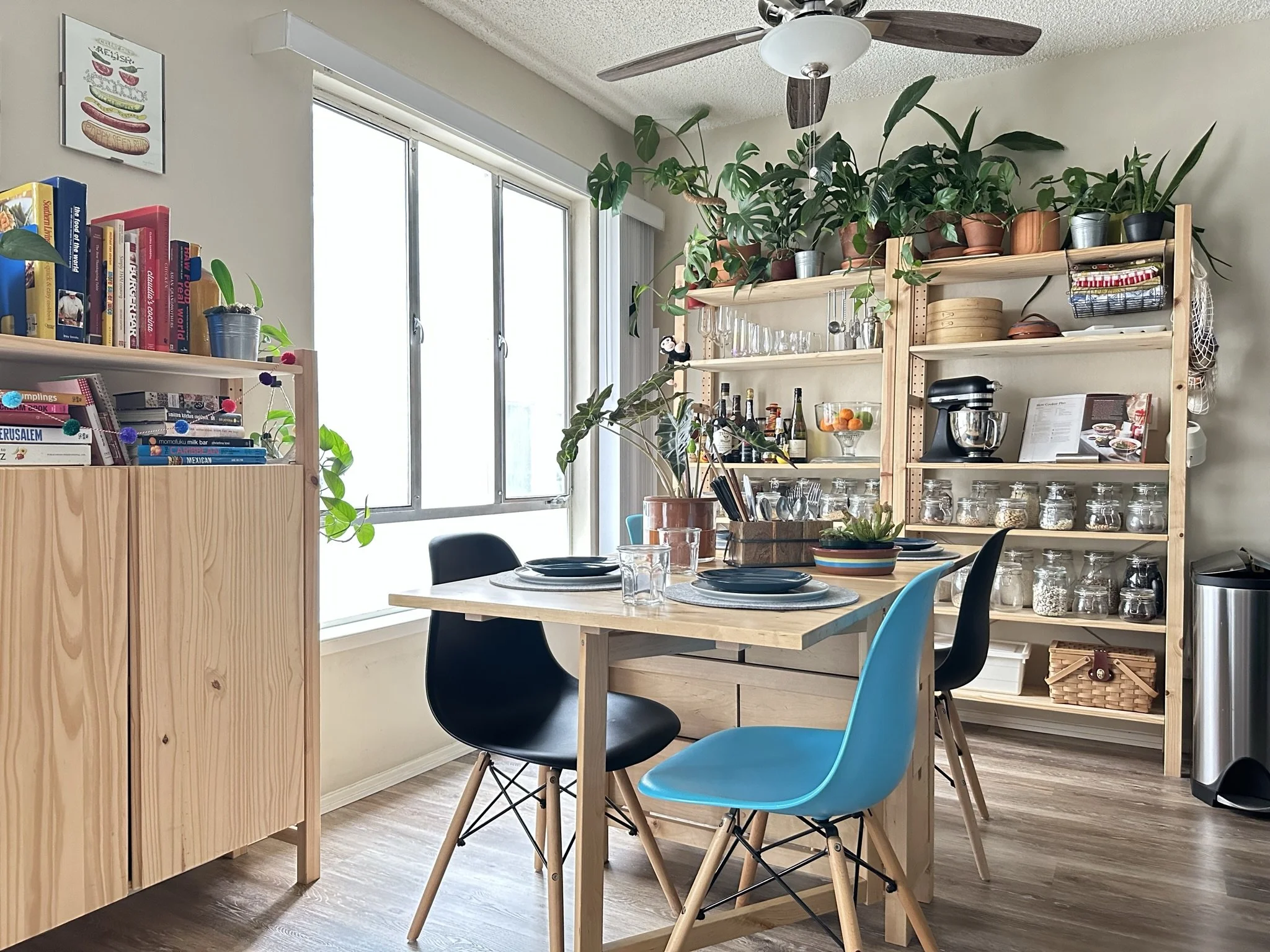 A cozy kitchen and dining area with a wooden table set with placemats and glassware, surrounded by colorful chairs, a large window providing natural light, and two wooden shelves filled with plants, glass jars, bottles, and kitchenware.