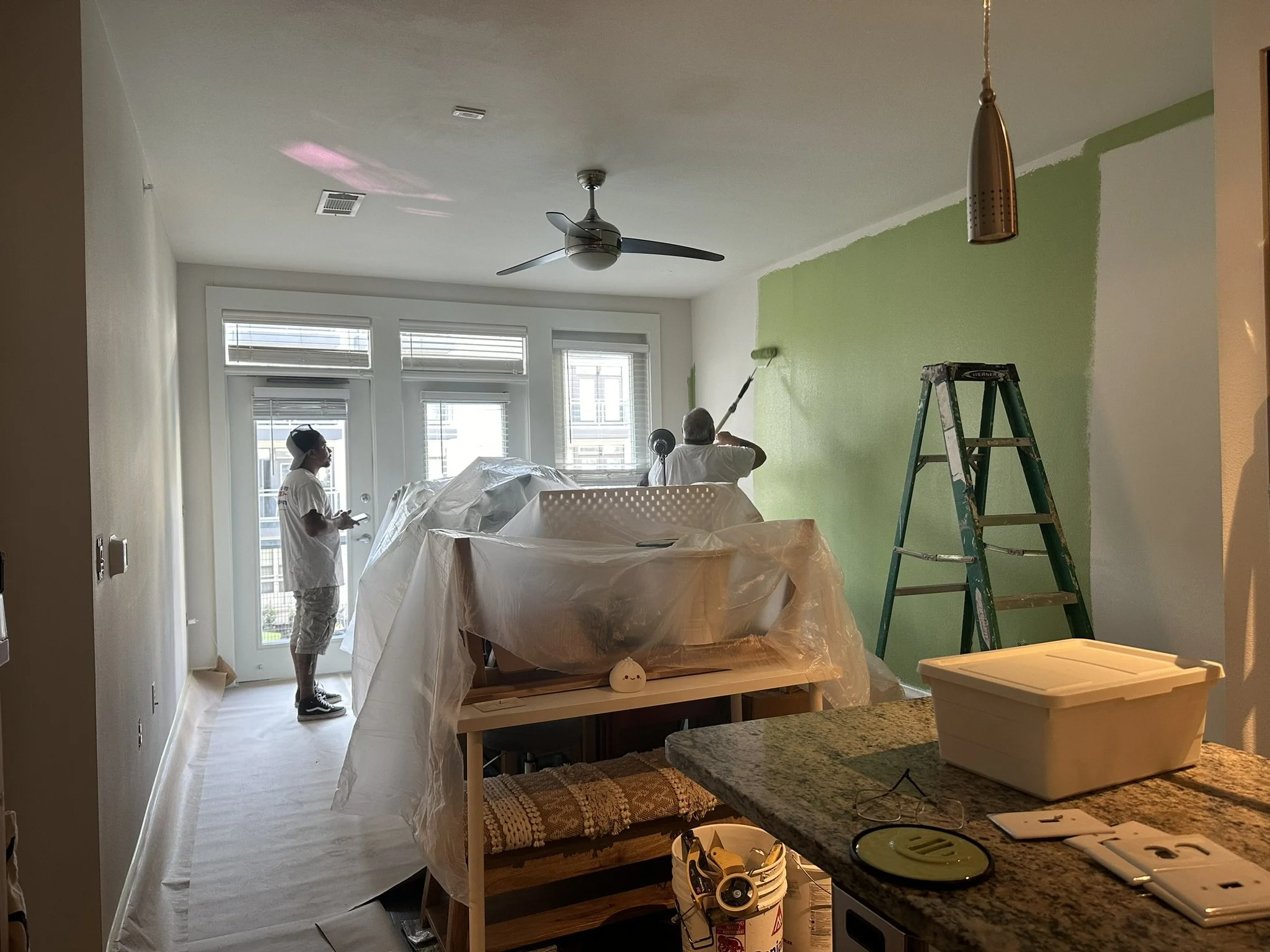 Two people painting a green wall in a living room, one painting with a roller and the other observing, with a ladder and painting tools nearby.