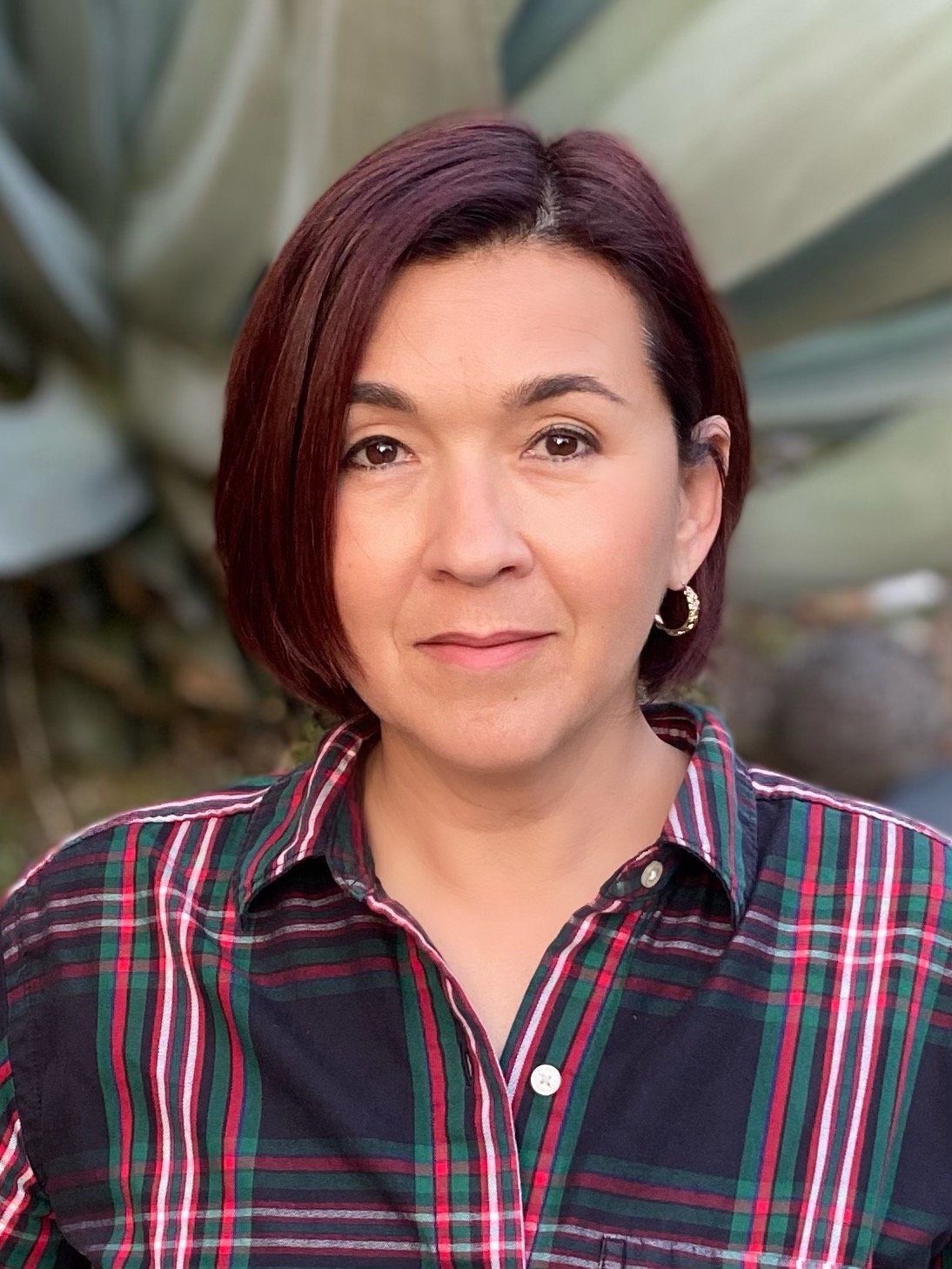 Person with short dark hair wearing a plaid shirt, standing in front of large green leaves.