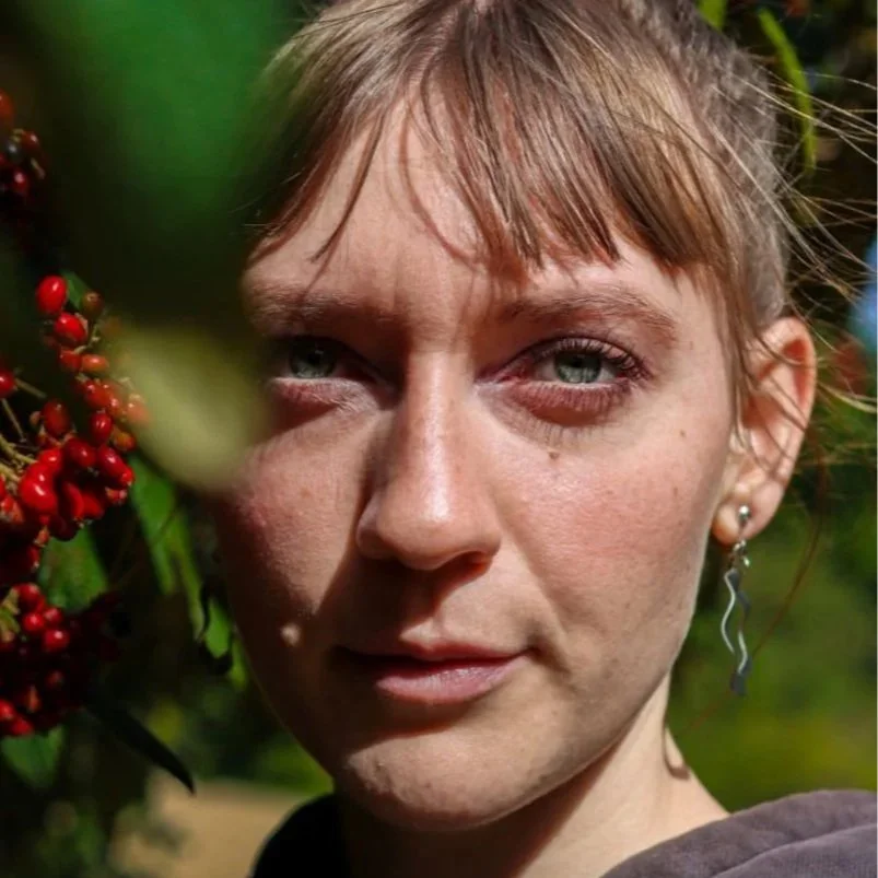 Nahaufnahme eines Mädchens mit blonden Haaren, blauen Augen und einem Ohrring, im Freien mit roten Beeren im Vordergrund.