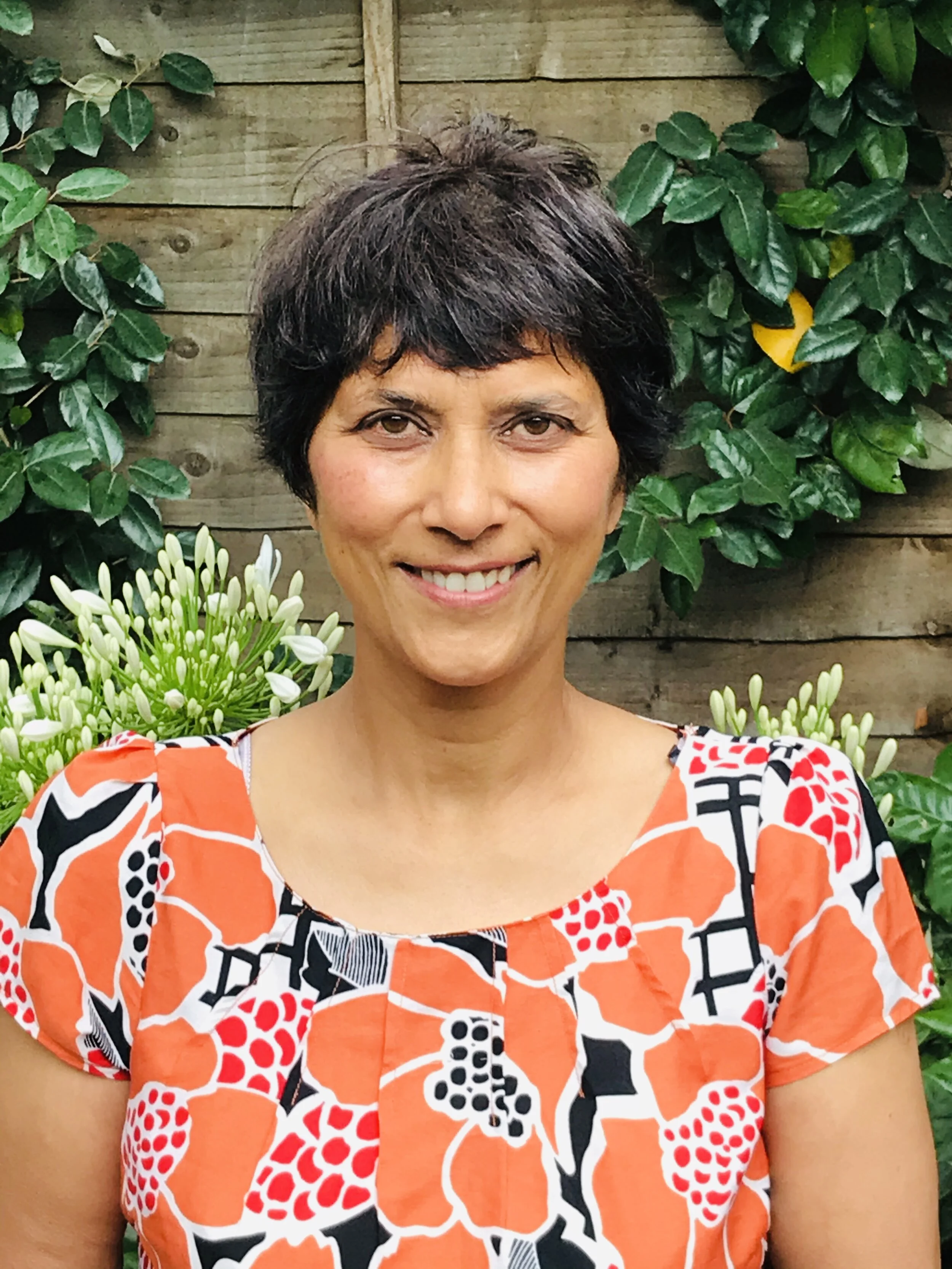 A woman with short dark hair smiling outdoors, standing in front of a wooden fence with green foliage and white flowers, wearing an orange, black, and white patterned top.