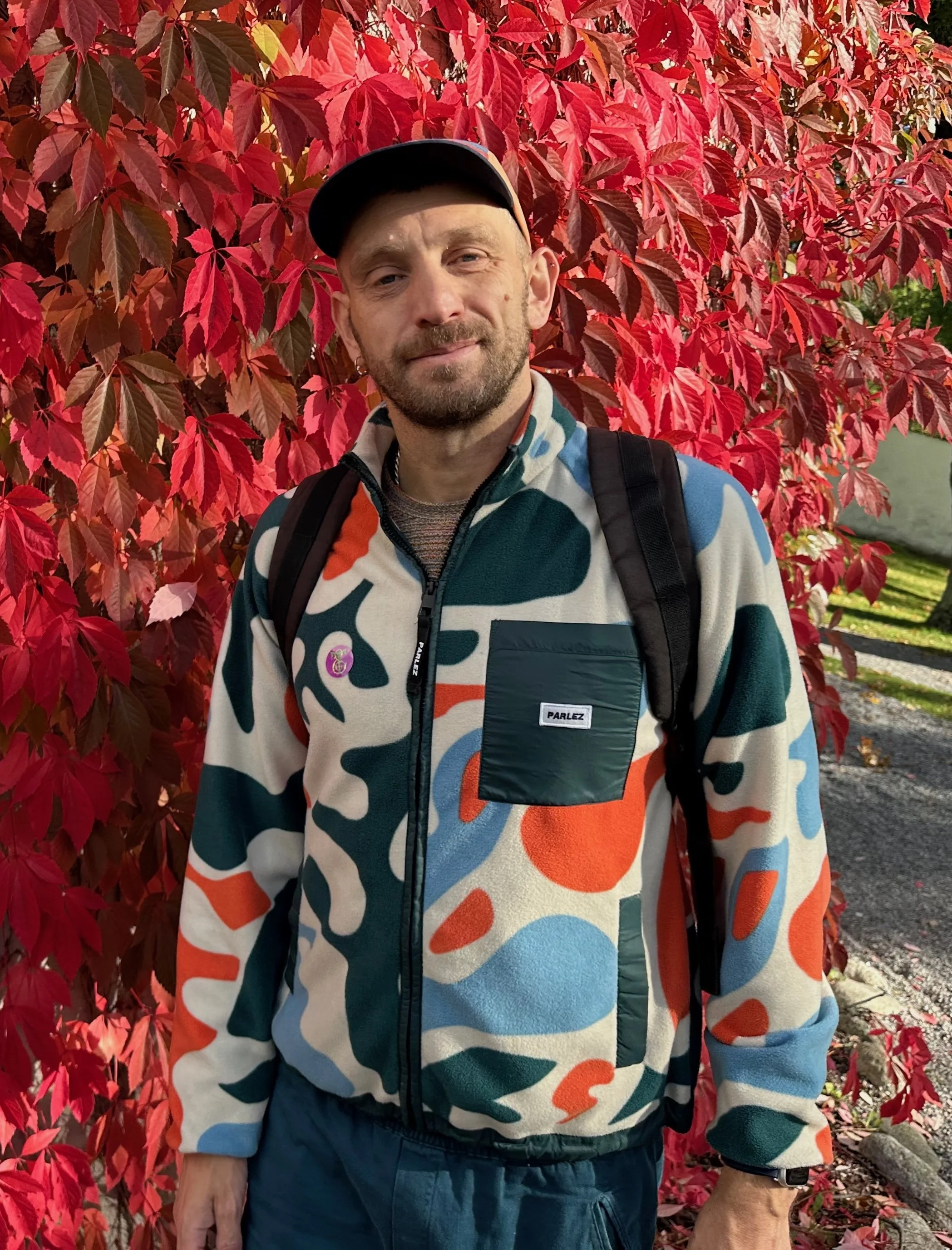 A man standing outdoors in front of a bush with red leaves, wearing a colorful patterned jacket, a black Cap, and a backpack.