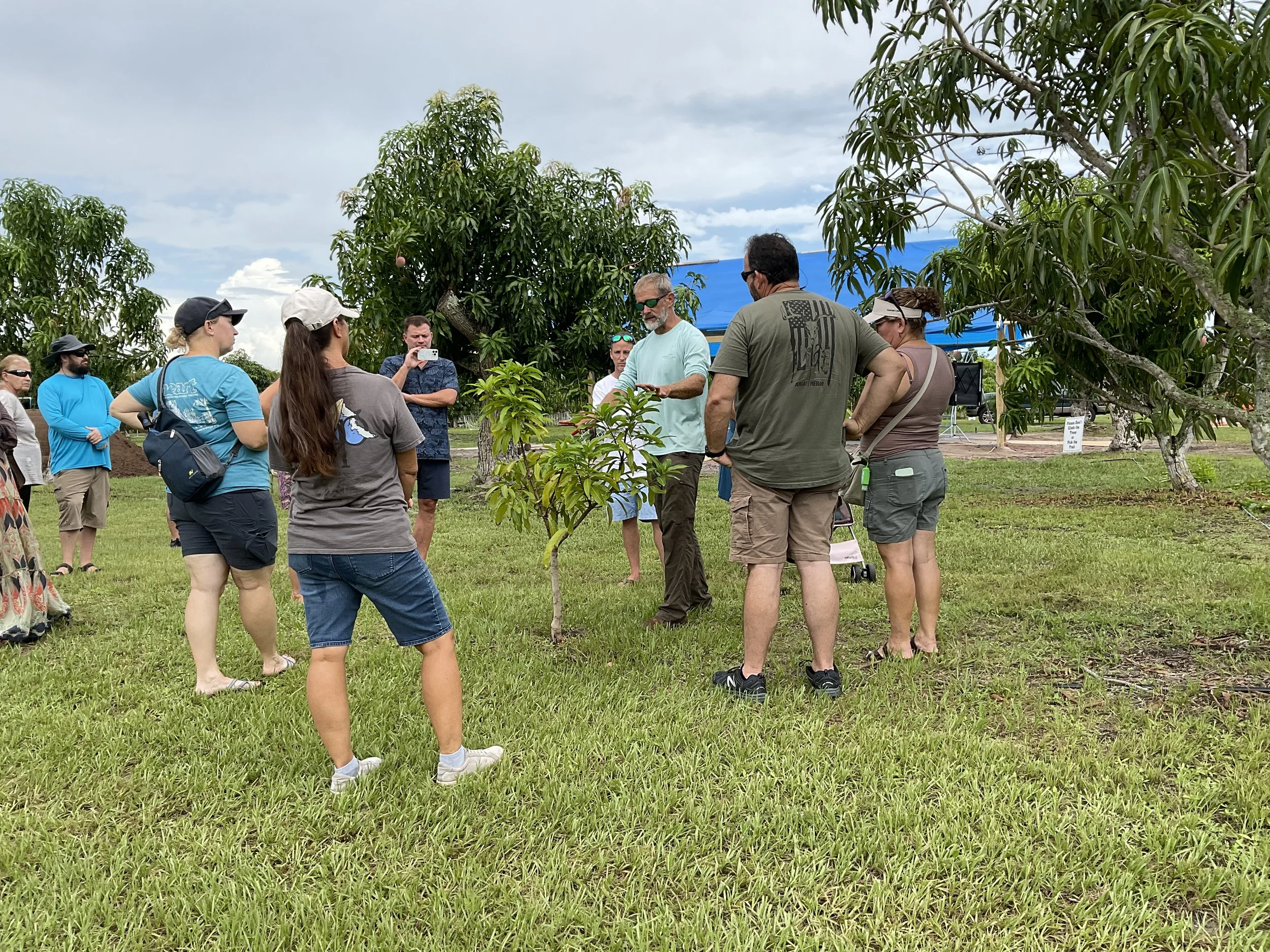 Group of people gathered outdoors, listening to a man speaking, with trees and a blue tent in the background.