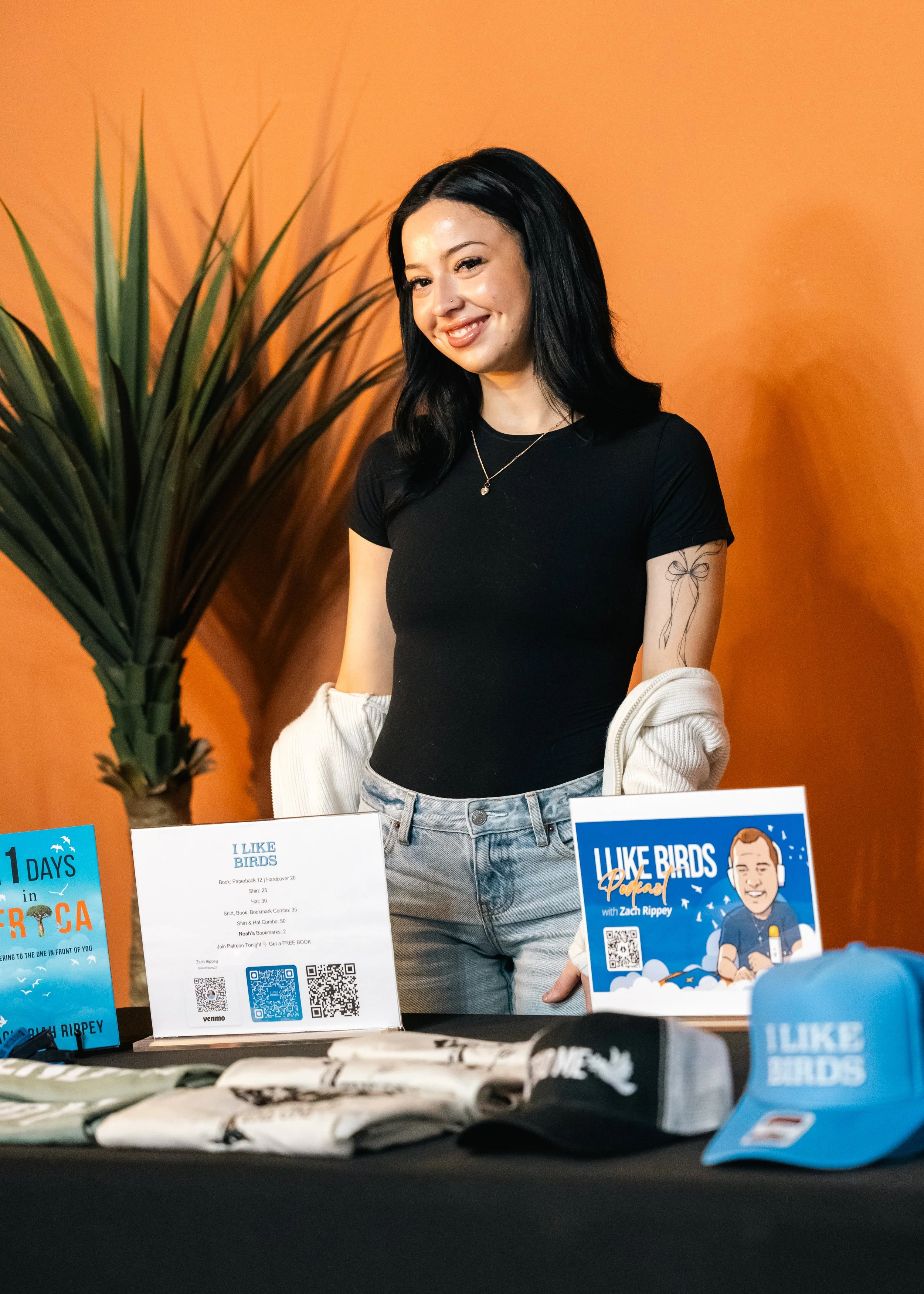 A smiling woman with black hair, wearing a black shirt and gray jeans, stands behind a table with promotional items, against an orange wall and a large green plant.