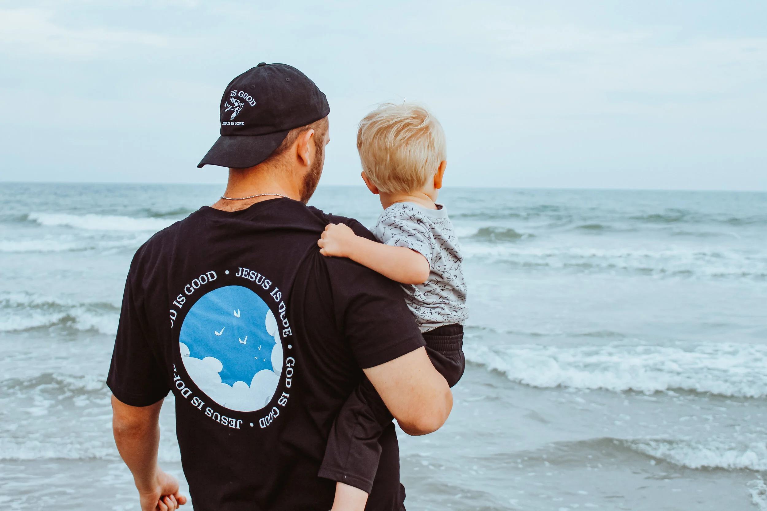 A man holding a young child on the beach, overlooking the ocean with waves and a cloudy sky in the background.