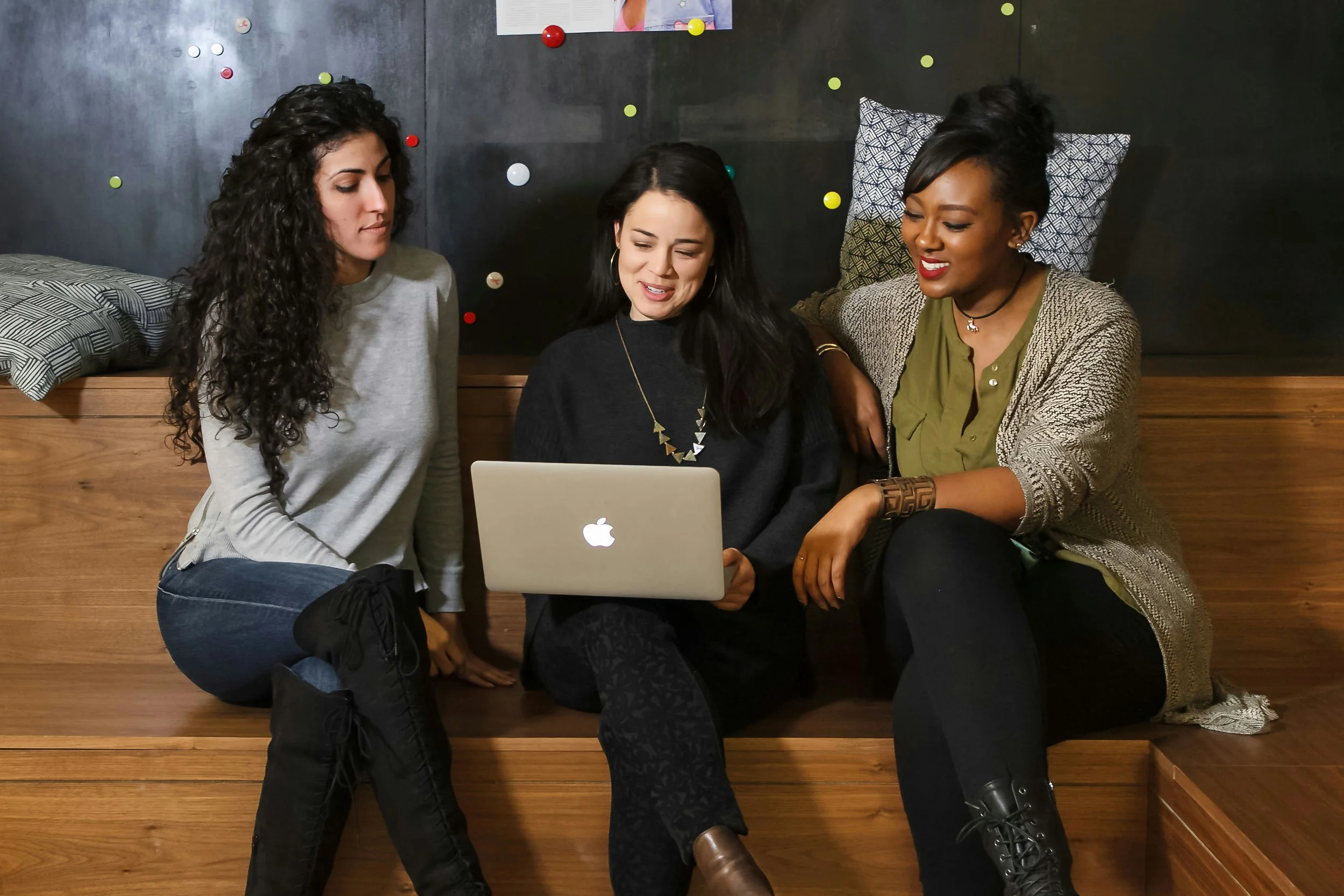 Three women sitting on a wooden bench, gathered around a laptop. They appear to be engaged and smiling, with a dark wall and colorful pins and a poster in the background.
