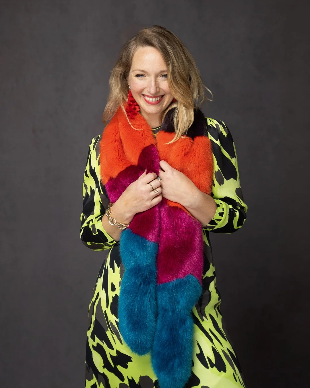 A smiling woman with blonde hair wearing a neon yellow and black patterned dress, holding a multicolored fur scarf with red, pink, and blue sections, against a dark gray background.