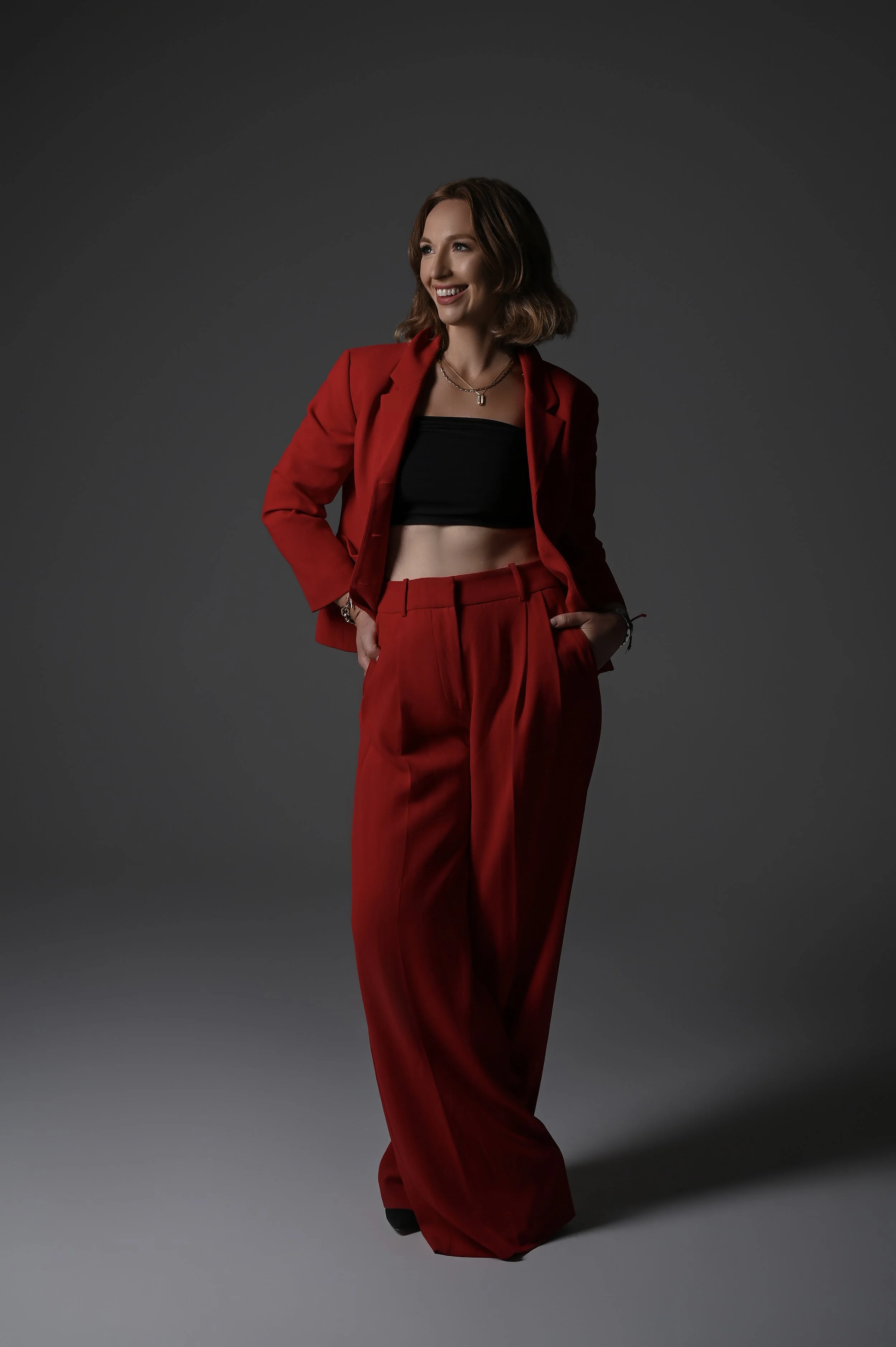 A woman with shoulder-length wavy brown hair, dressed in a red blazer and matching red trousers, smiling and standing with her hands in her pockets against a gray background.