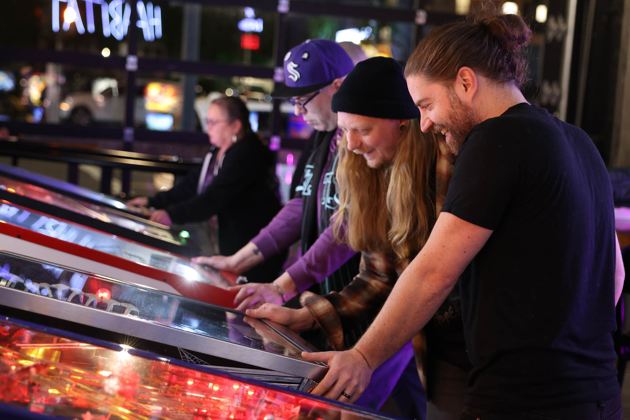 Four people play pinball at an arcade, smiling and enjoying the game.