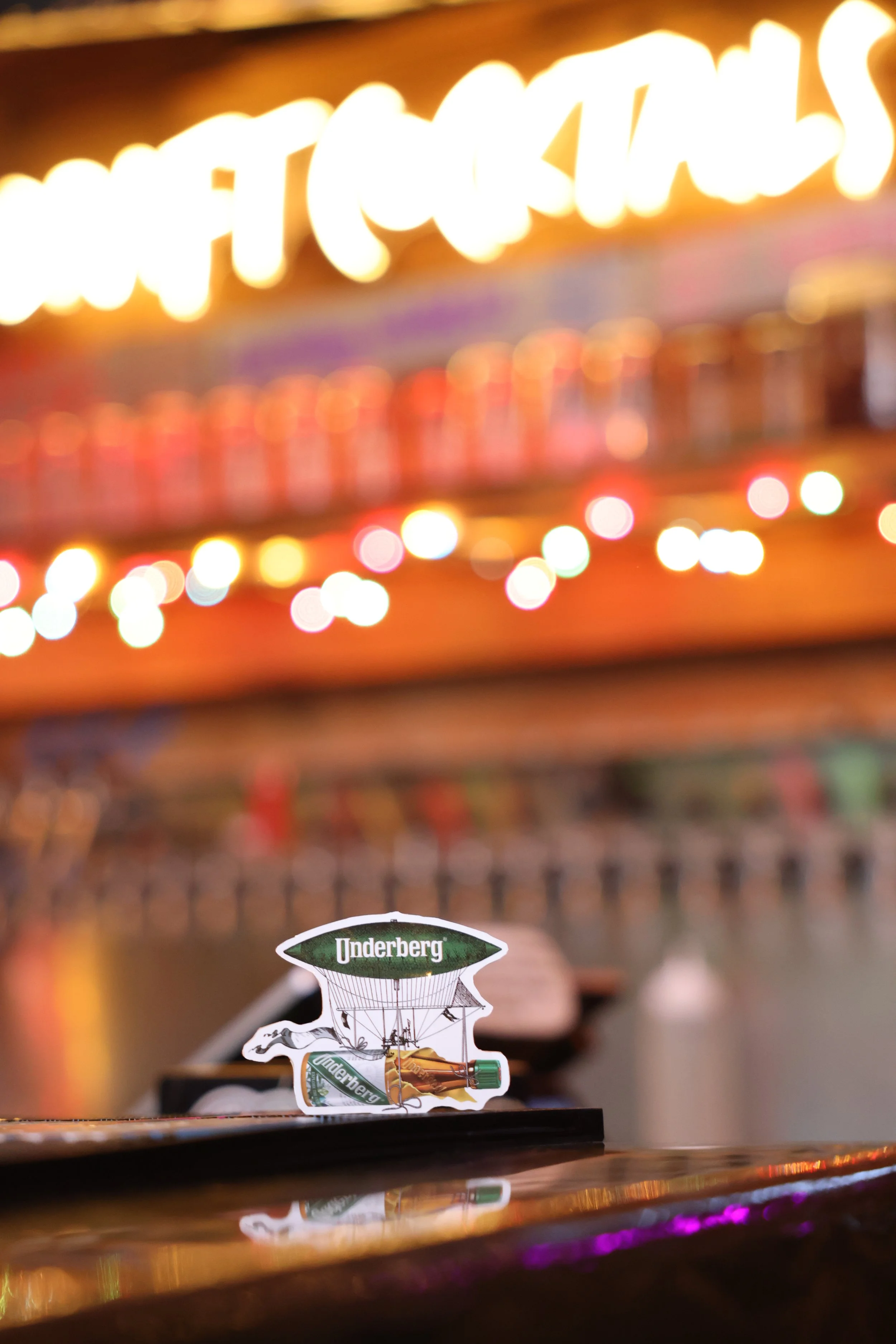 A small paper cutout of a hot air balloon with the word 'Underberg' on the balloon part, placed on a bar counter with colorful lights and blurred background.