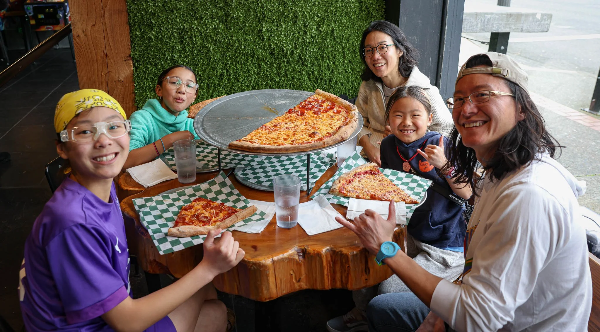 A group of six people, including three children and three adults, sitting around a wooden table in a pizzeria, enjoying pizza. The table has two large pizza slices and a whole pizza on a hot plate. Everyone is smiling and having a good time.