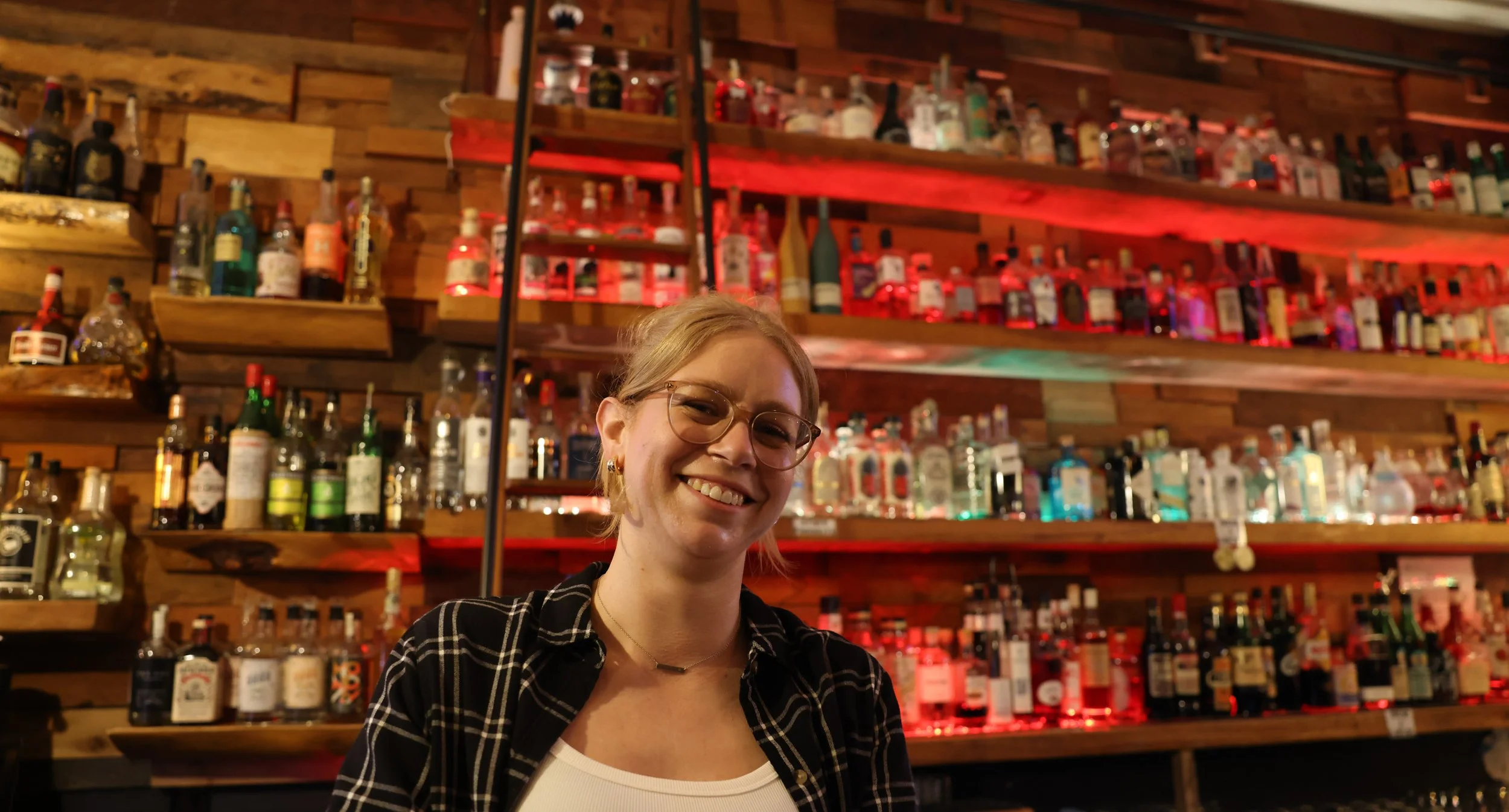 A smiling woman with glasses and earrings, wearing a black plaid shirt over a white top, standing in front of a wooden bar with various bottles of alcohol on shelves behind her.