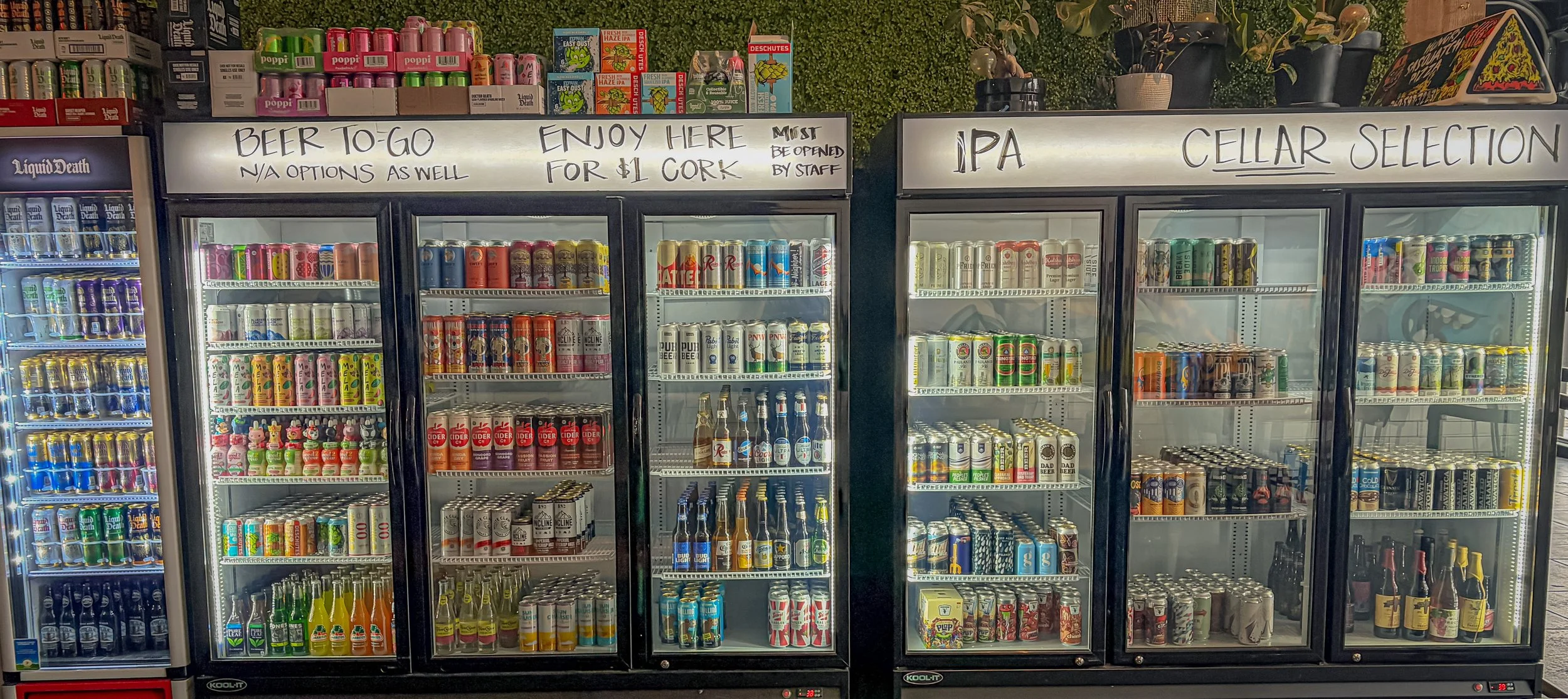 Refrigerators filled with various beers in a liquor store, with handwritten signs above indicating options like 'Beer To-Go,' 'Enjoy Here,' and 'Cellar Selection'.