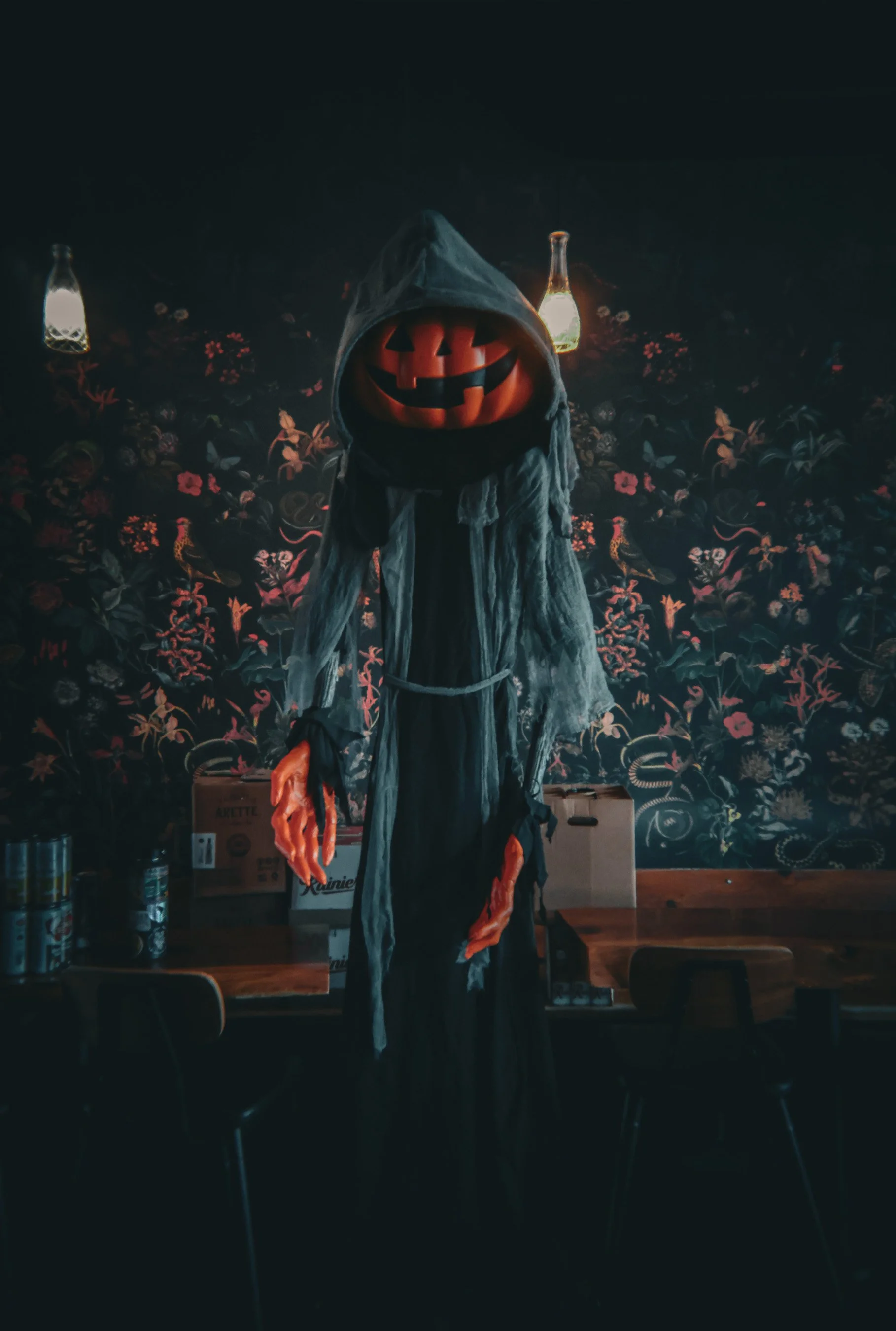 A Halloween decoration of a spooky figure with a pumpkin head, dressed in tattered gray and black robes with orange gloves, standing in a dimly lit room with floral wallpaper and hanging lanterns.