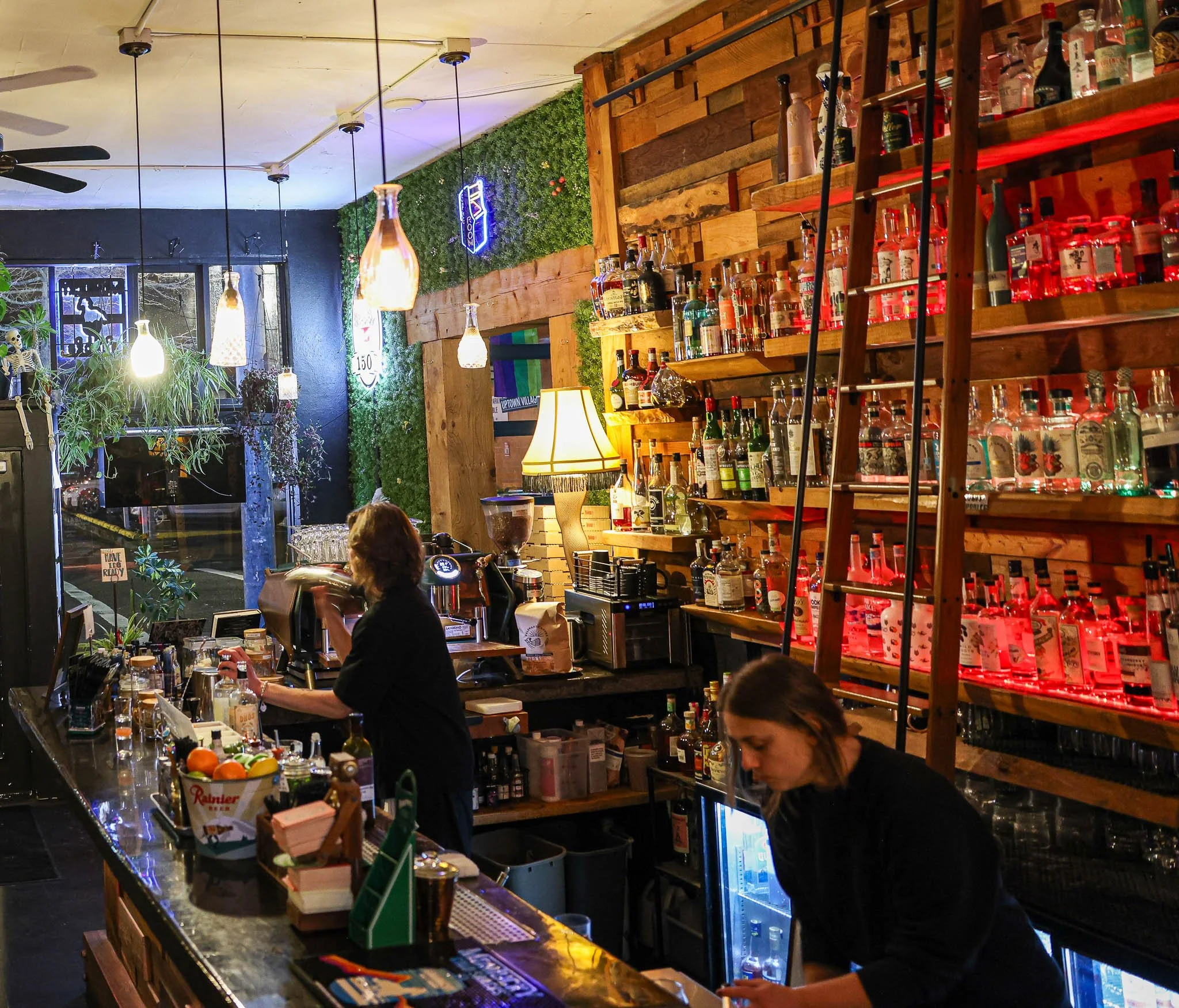 A cozy bar with a bartender preparing a drink and a staff member working. Shelves behind the bar hold various bottles of alcohol, and there's a warm yellow lamp. The interior includes wood accents, a green plant, and string lights.