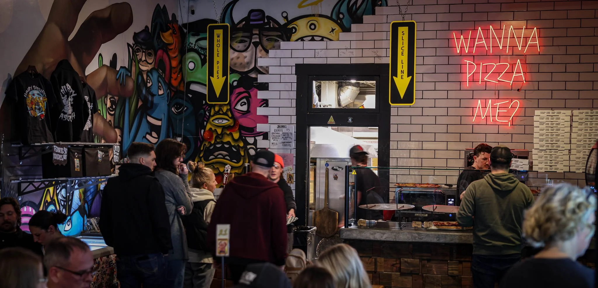 Inside a busy pizzeria with colorful graffiti art on the wall, neon sign reading 'Wanna Pizza Me?' on the brick wall, and customers waiting in line.