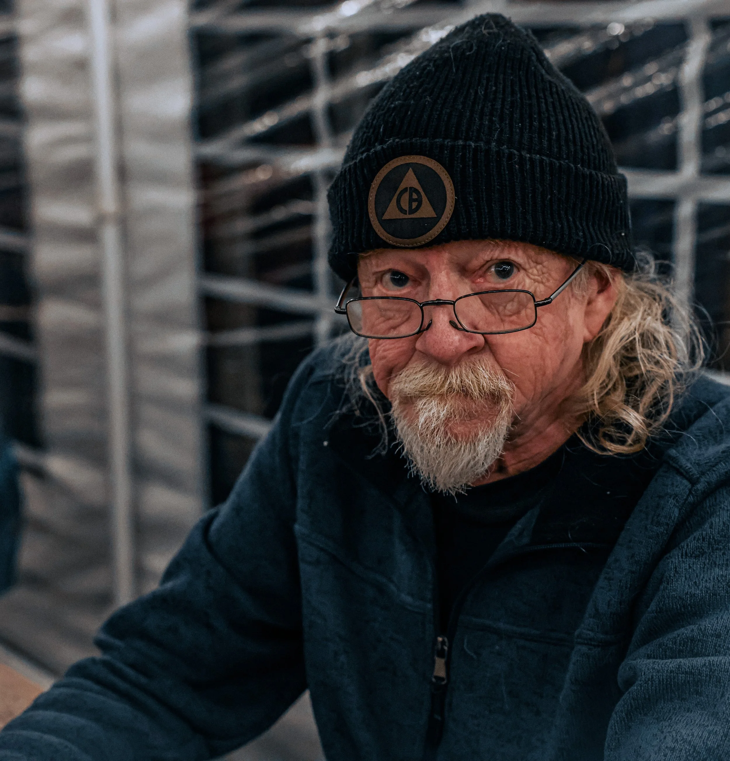 An older man with long gray hair and beard wearing glasses, a black beanie with a logo, and a dark jacket, sitting in front of a metal rack with shelves.