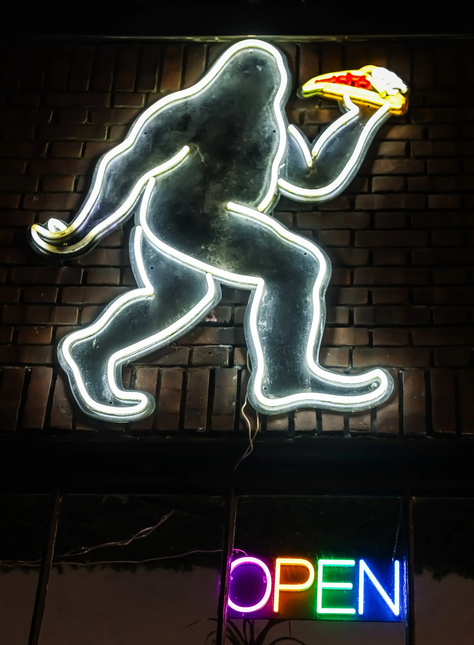 Neon sign of a muscular person holding a pizza, with a smaller neon sign below that says 'OPEN' in rainbow colors.